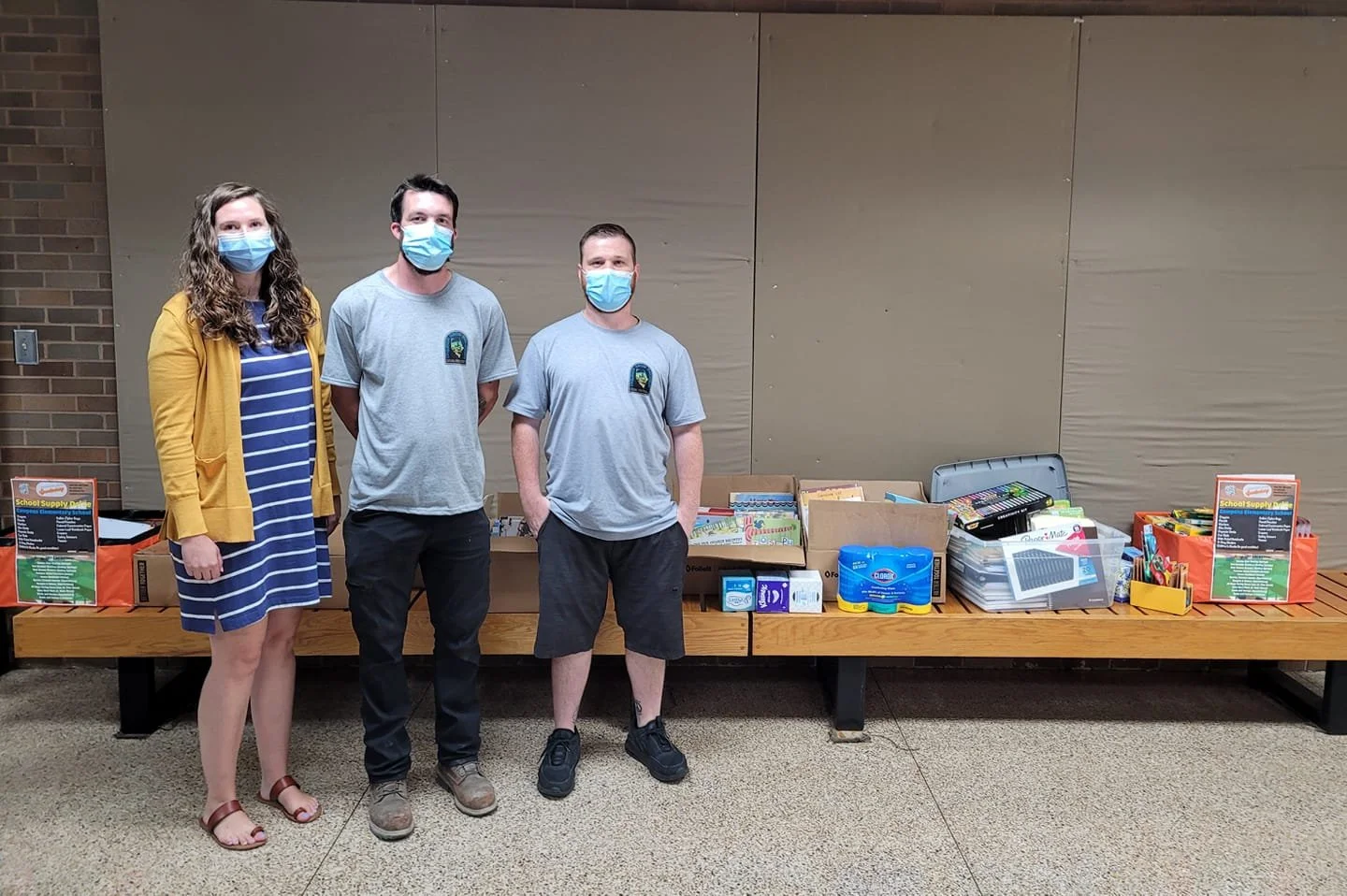 Three people wearing masks standing in front of a table filled with school supplies including boxes, notebooks, and art supplies.