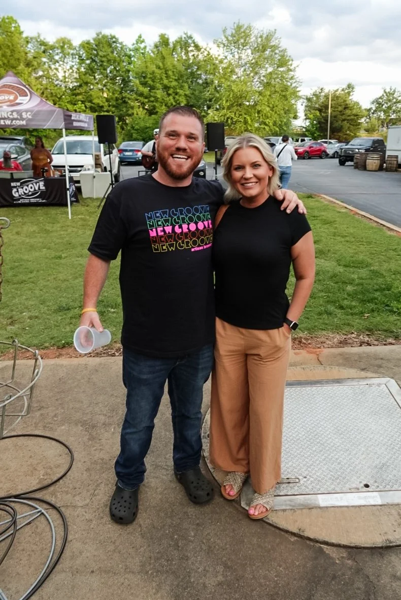A smiling man and woman standing outdoors on a sidewalk with greenery and parking lot in the background, posing together with the man's arm around the woman's shoulder.