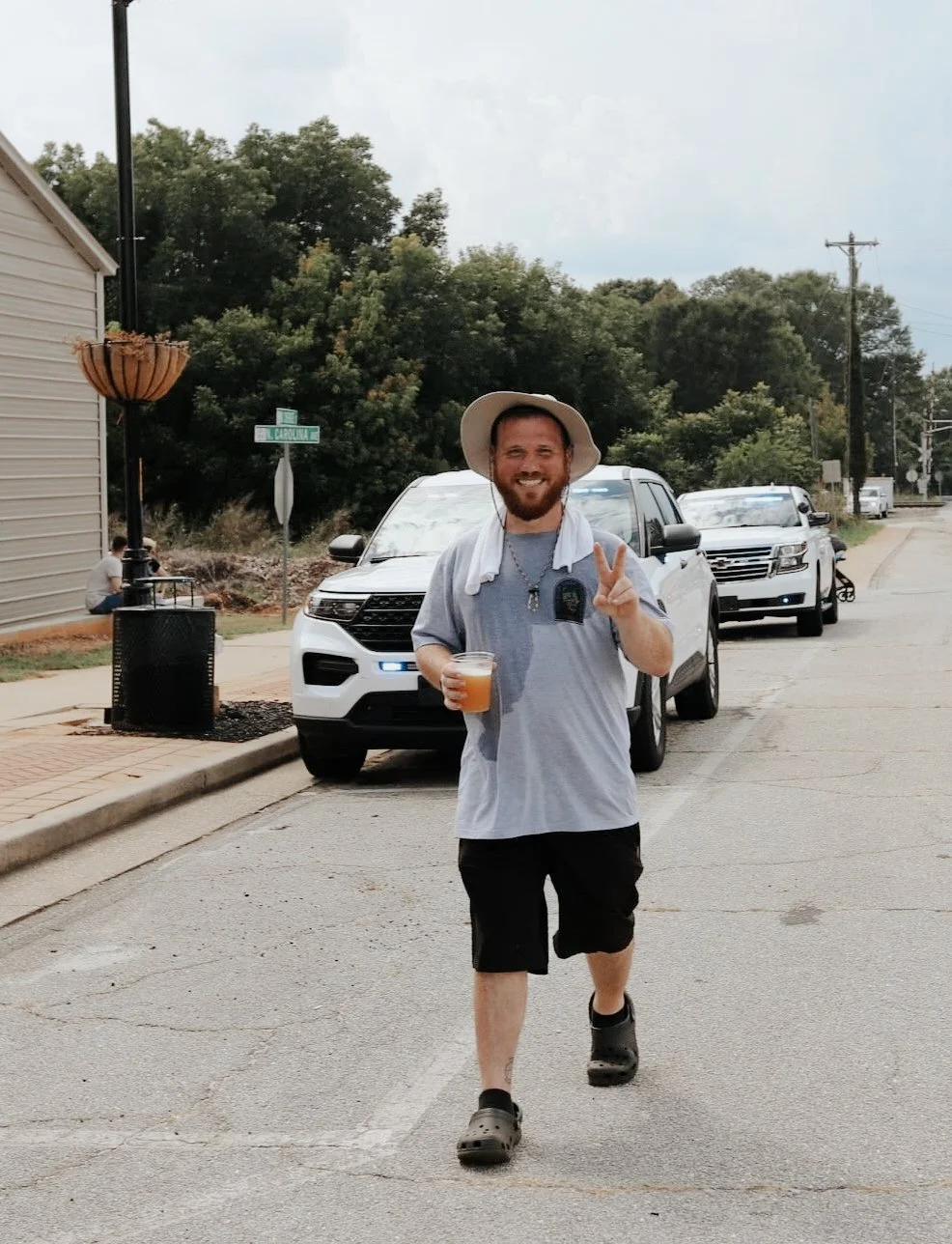 A man with a beard, wearing a wide-brimmed hat, gray T-shirt, black shorts, and Crocs, is smiling and making a peace sign while holding a drink. He is walking on a street with parked cars, a sidewalk, trees, and a street sign in the background.