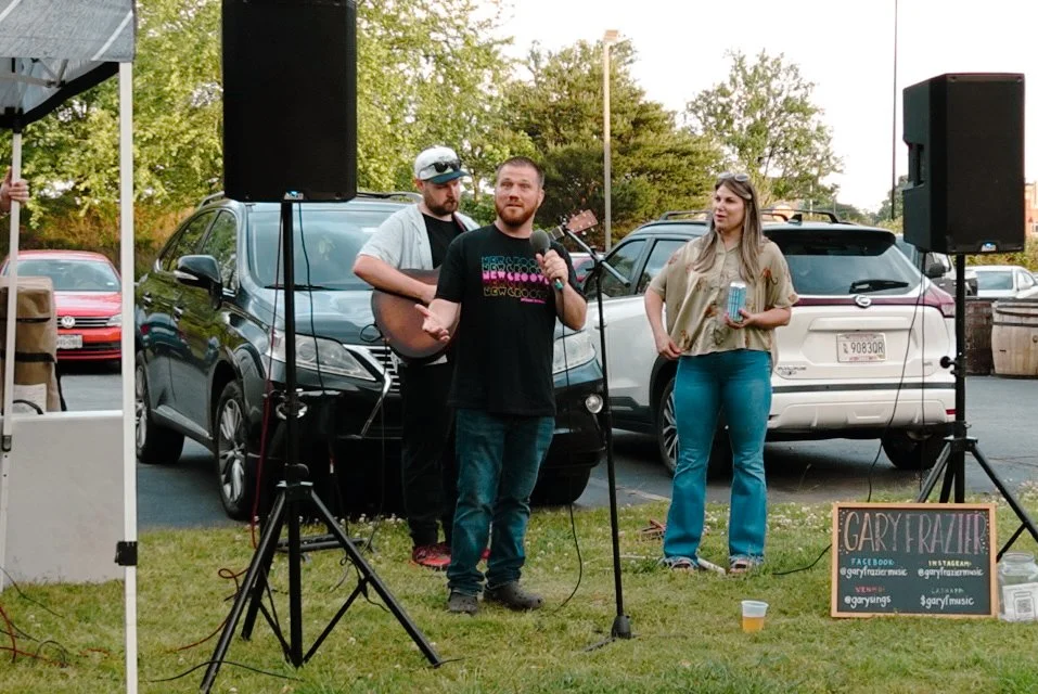 A man with a beard and glasses is speaking into a microphone while two people, a man with a guitar and a woman holding a drink, stand in the background. They are outdoors near parked cars, with trees in the background and two large speakers set up on either side of the speakers.