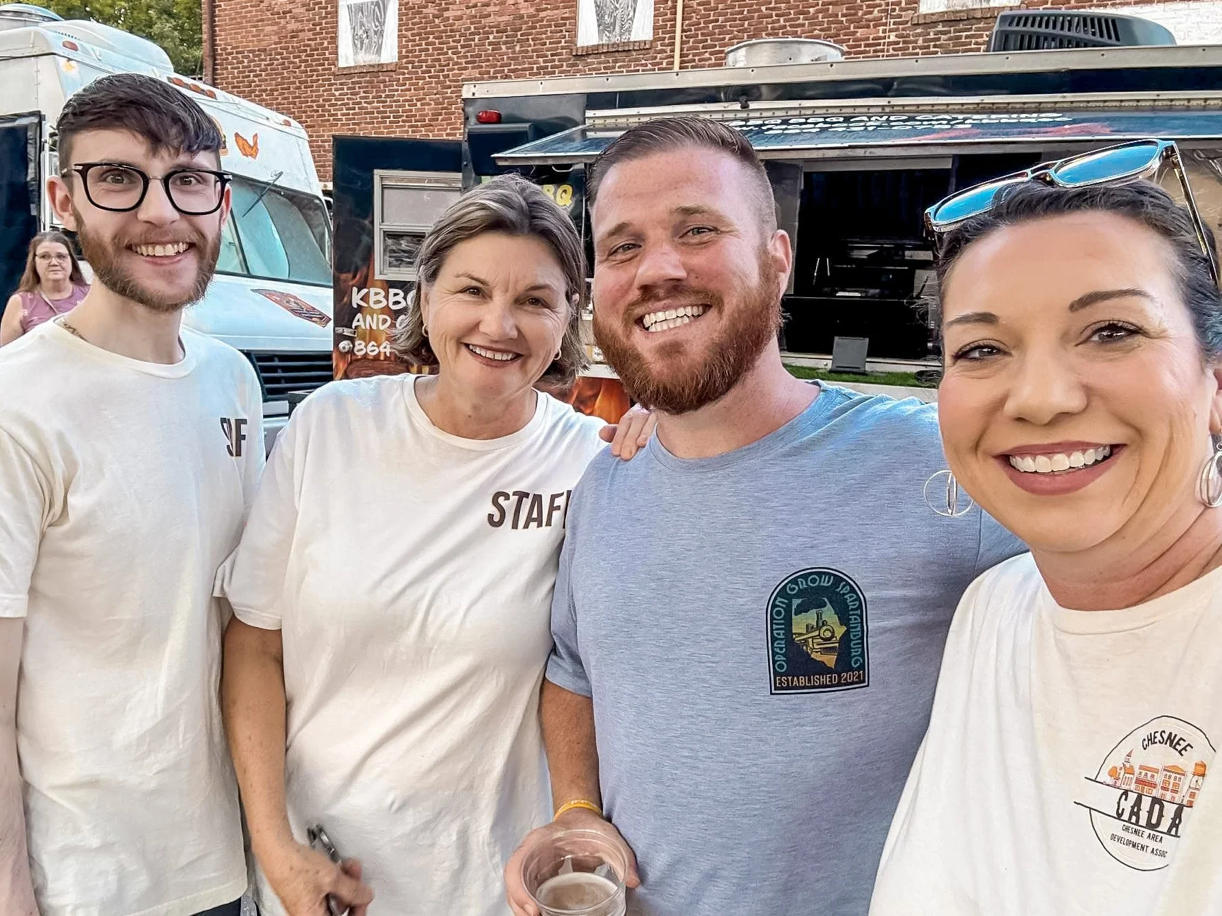 Group of four people smiling in front of food trucks at an outdoor event, three women and one man, during daytime.