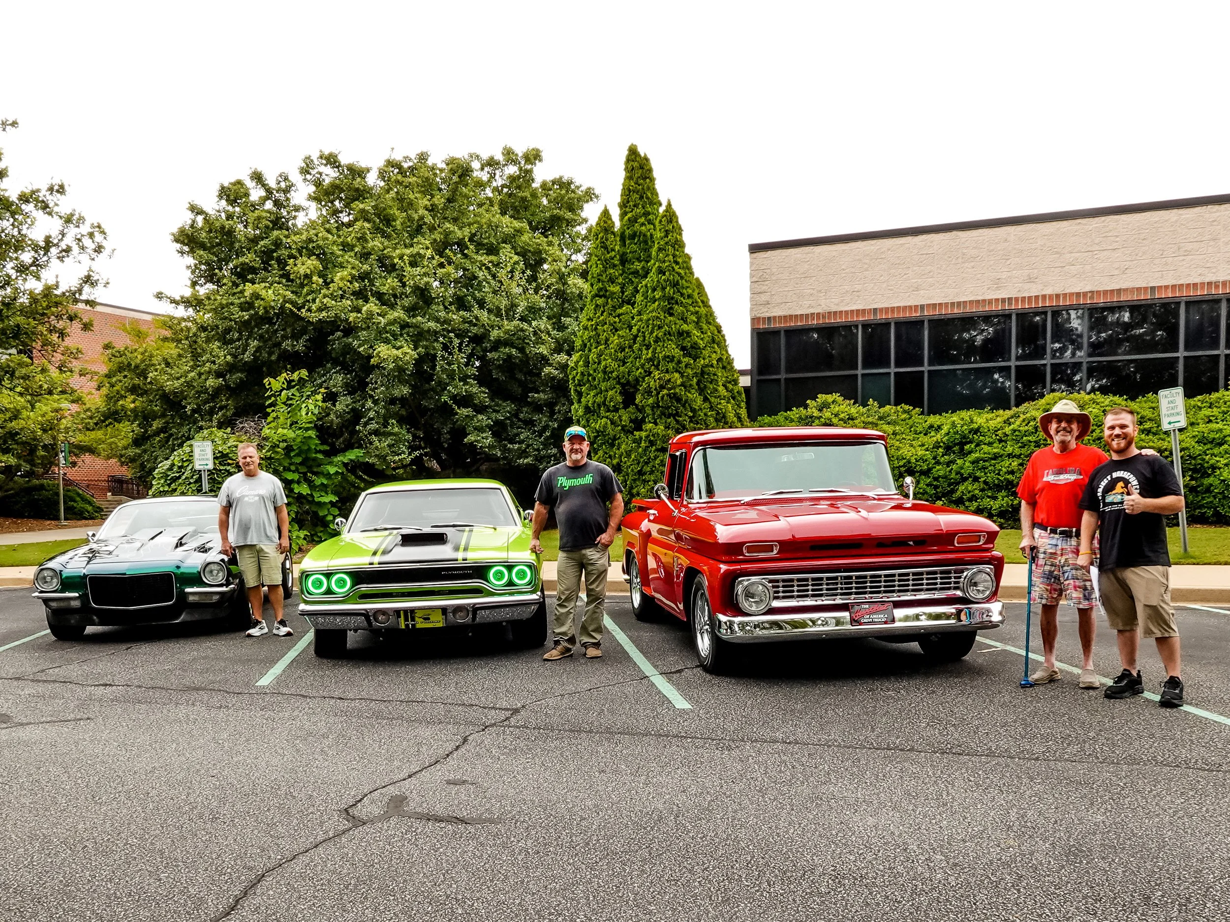 Four men standing next to three classic cars in a parking lot, with trees and a building in the background.