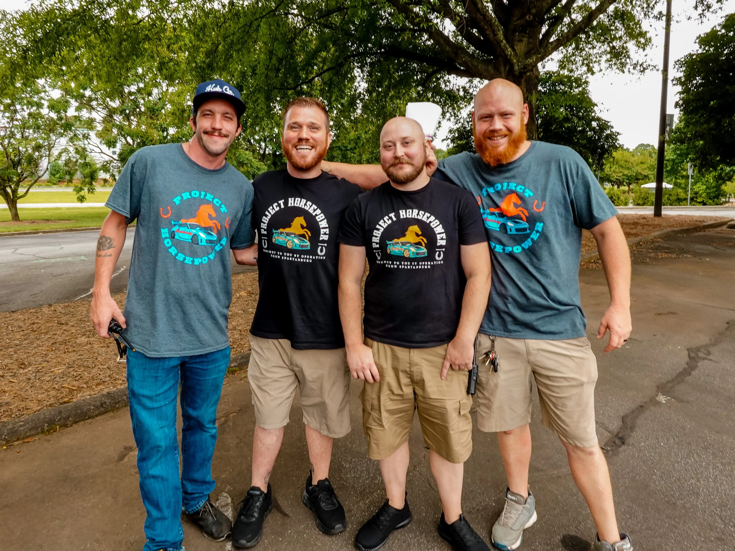 Four men standing outdoors on a parking lot with trees in the background, smiling and posing for the photo, wearing T-shirts with a car and horse logo.
