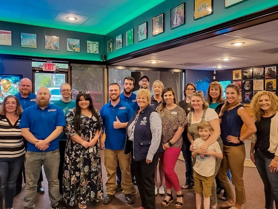 Group of people indoors, some wearing blue shirts, with a woman holding a young boy, in front of a wall decorated with artwork.