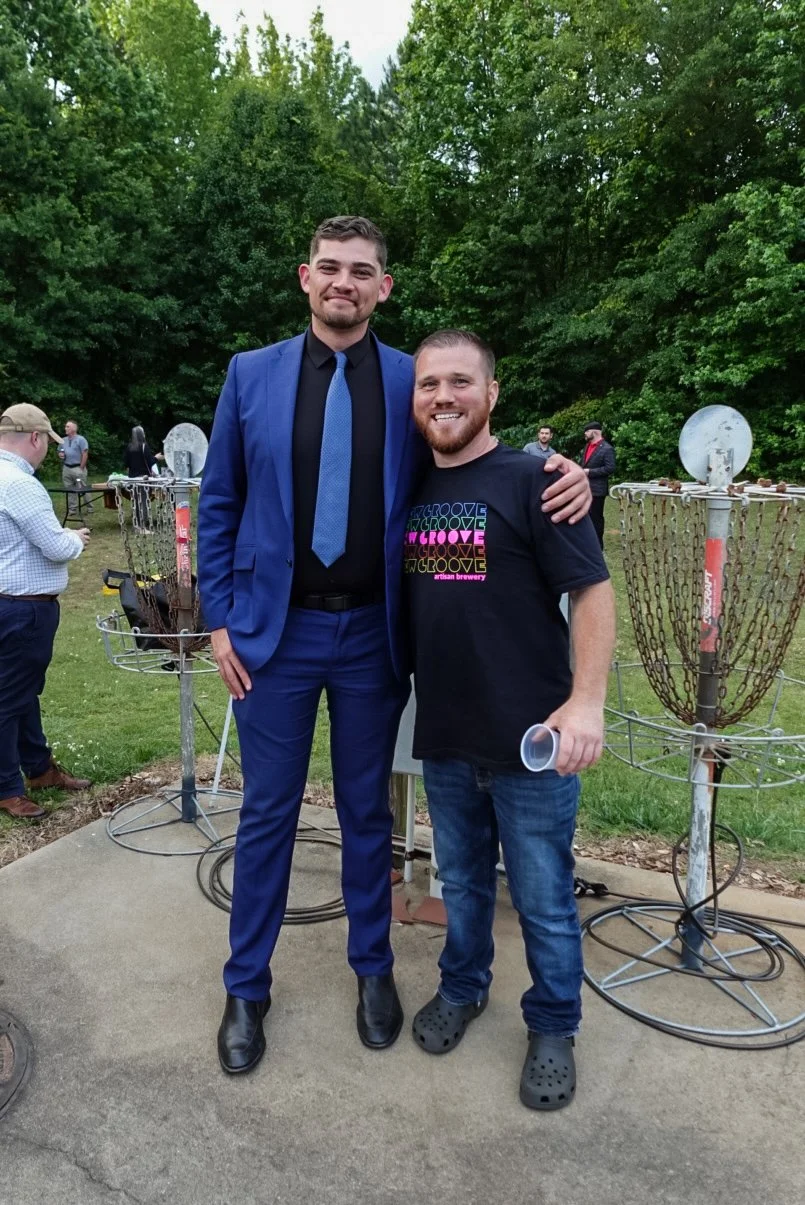 Two men standing outdoors among trees, one dressed in a blue suit and the other in a black graphic t-shirt and jeans, smiling and posing together. There are disc golf baskets and other people in the background.