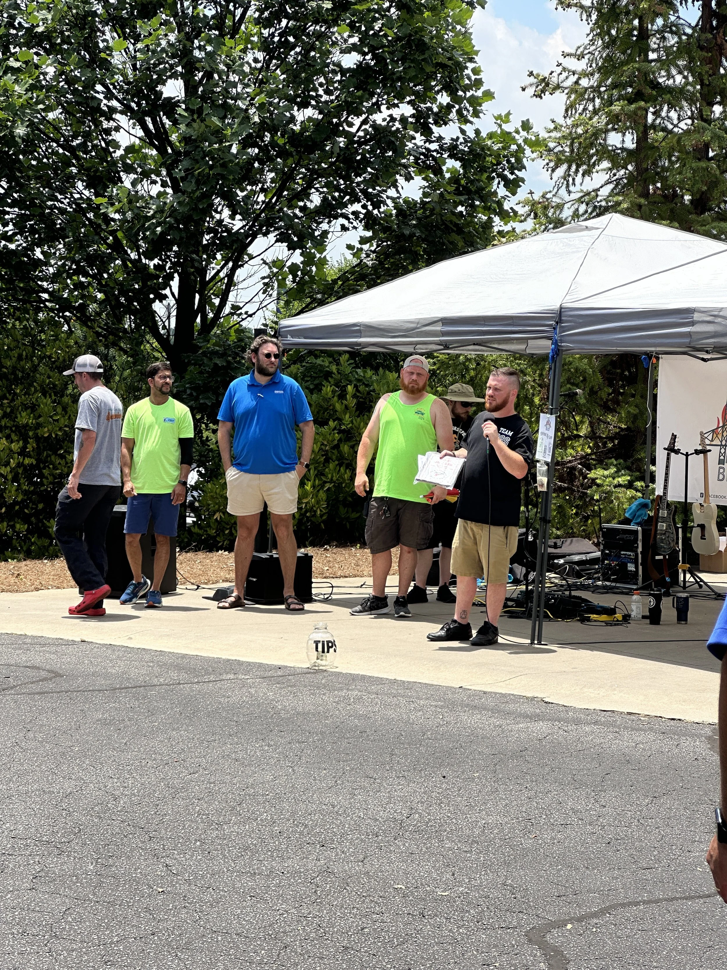 A group of six men standing under a canopy on a sidewalk during daytime. One man is speaking into a microphone. The men are dressed casually in shorts and T-shirts, with some wearing bright green shirts. There are trees and greenery in the background.