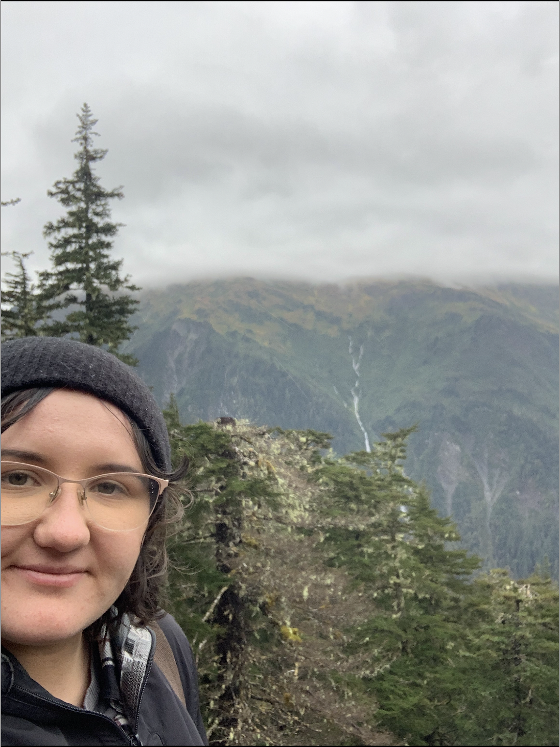 A person smiling with glasses and a beanie hat, standing in a forested mountain area with mountains, trees, and a waterfall in the background under a cloudy sky.