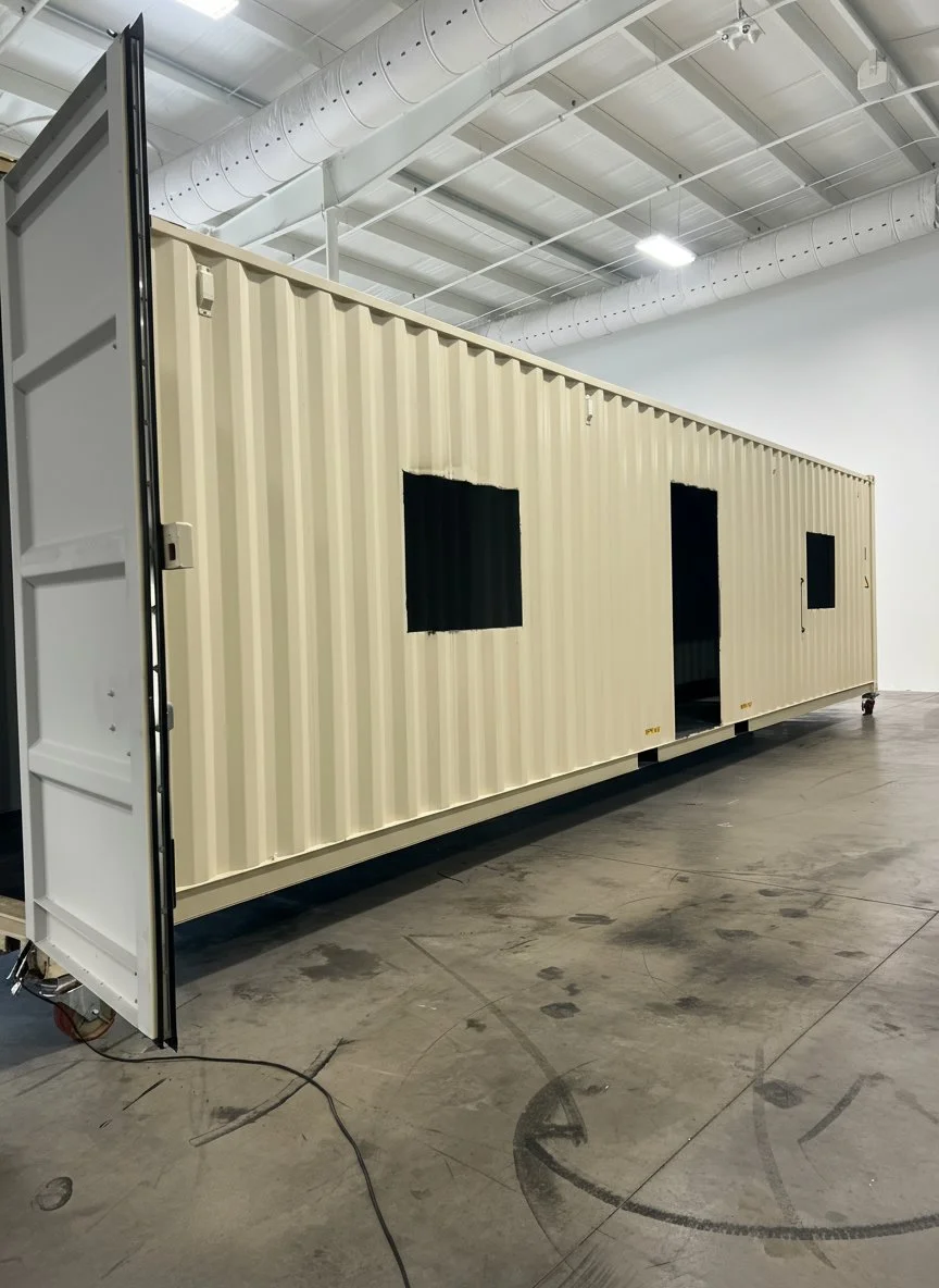 A large beige shipping container with windows and an open door, placed inside an industrial space with a concrete floor and white ceiling with ductwork.