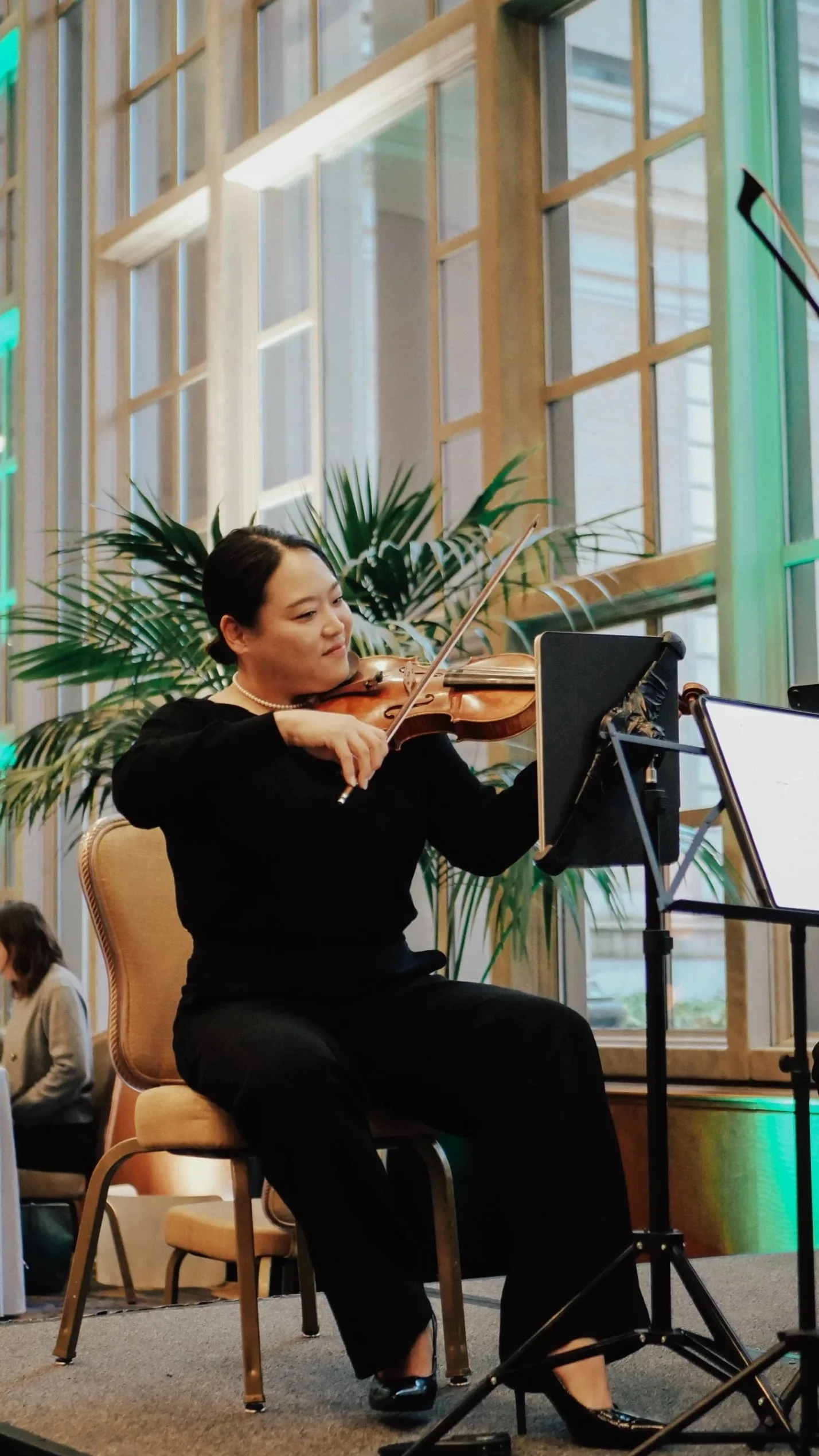 A woman playing violin during a performance indoors with large windows and greenery in the background.