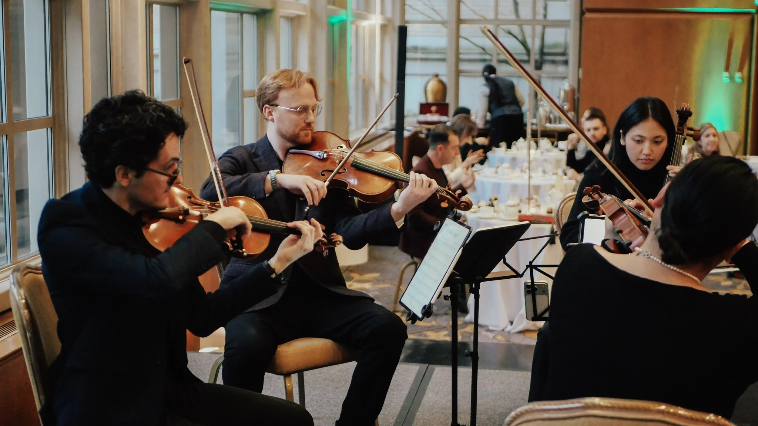 A group of four musicians playing violins during a performance at a formal event, with a decorated dining area in the background.