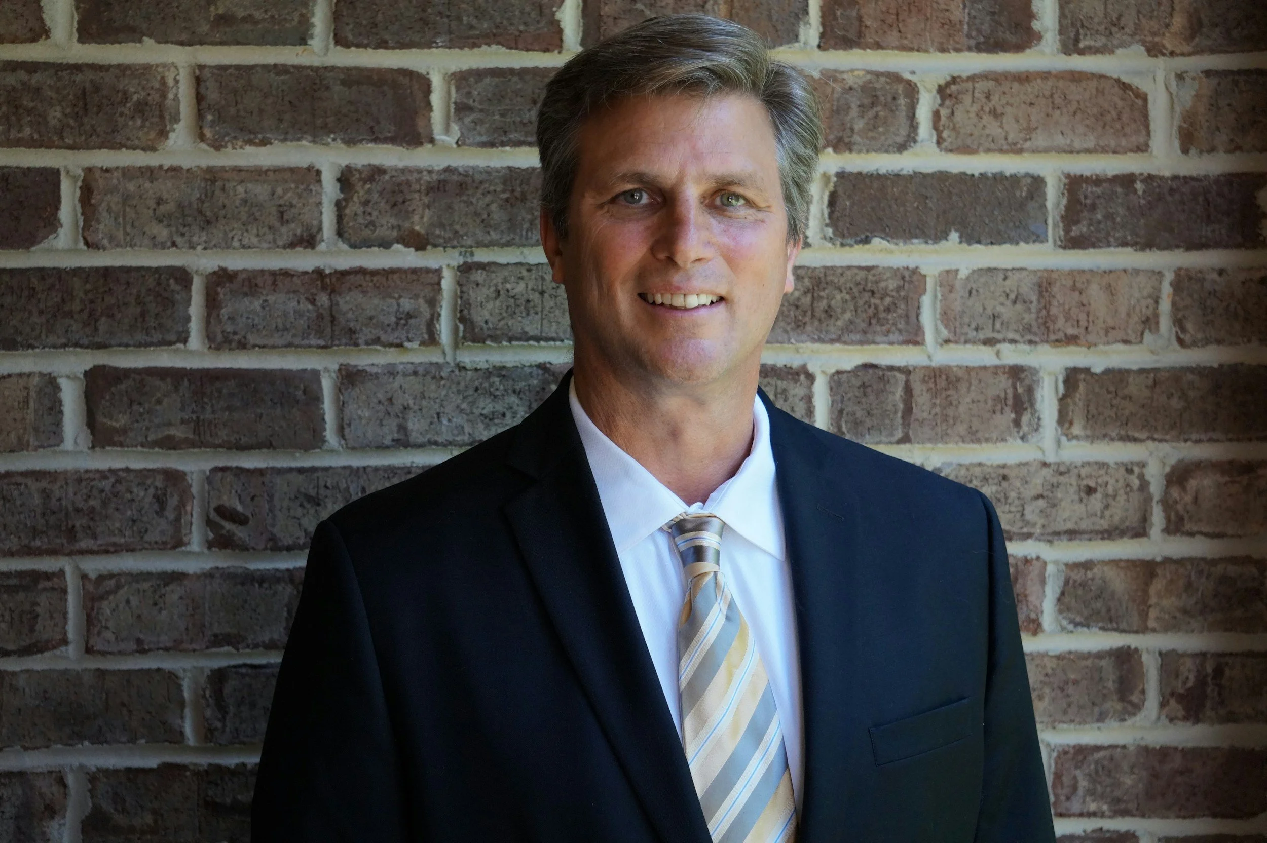 A middle-aged man with light skin, gray hair, and blue eyes, smiling, wearing a dark suit, white shirt, and a yellow striped tie, standing in front of a brick wall.