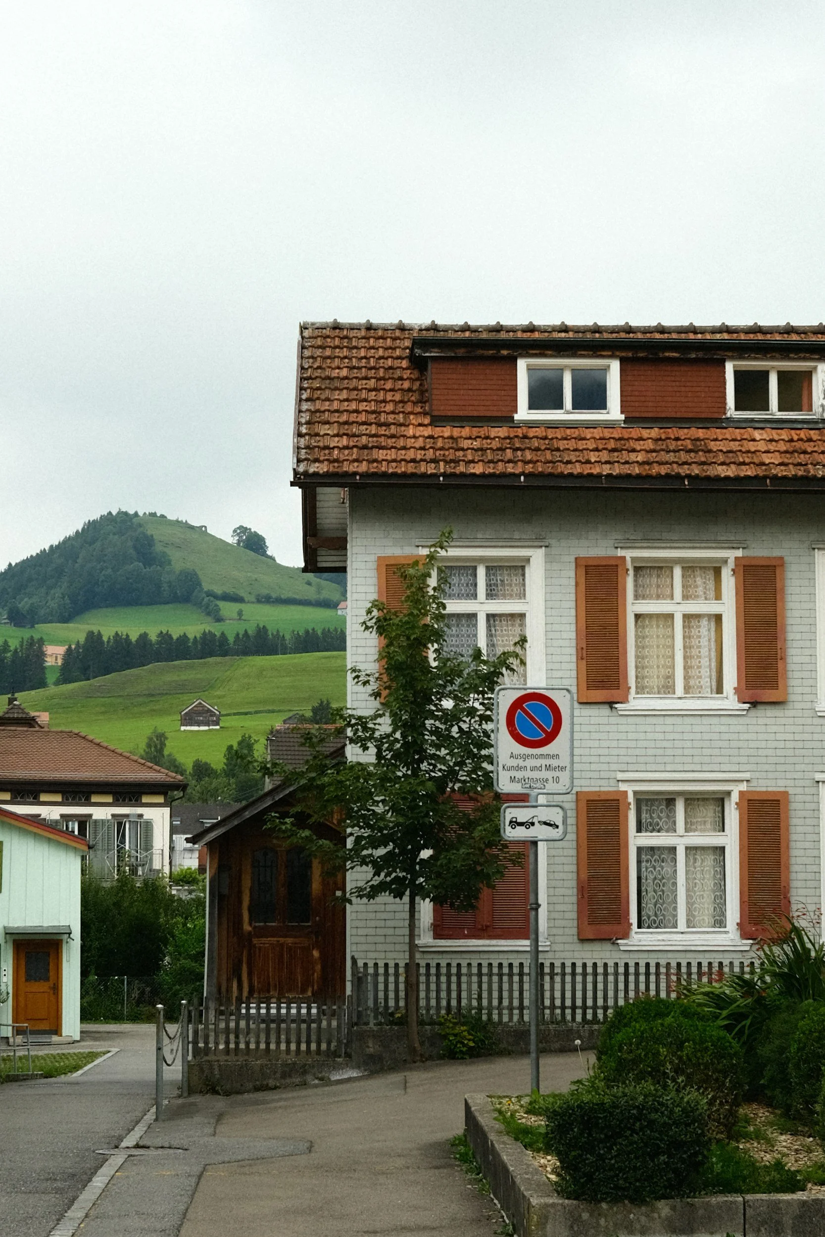 A white house with brown window shutters and a red tiled roof, a small tree in front, and a no parking sign in German on a street with green hills in the background.