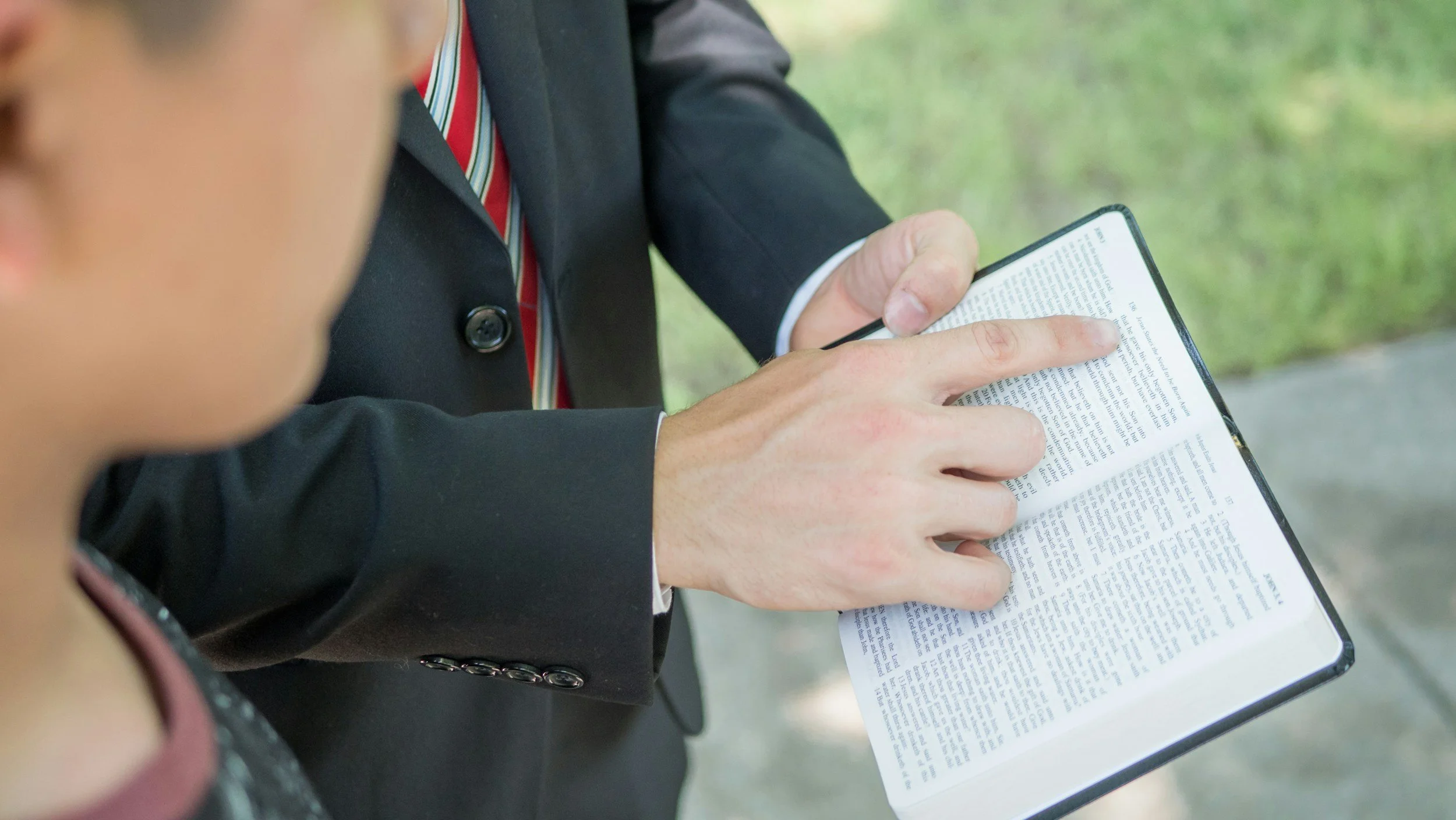 A person in a dark suit with a striped red, green, and white tie is holding and reading a small open book or pamphlet outdoors on a paved path, with grass in the background.