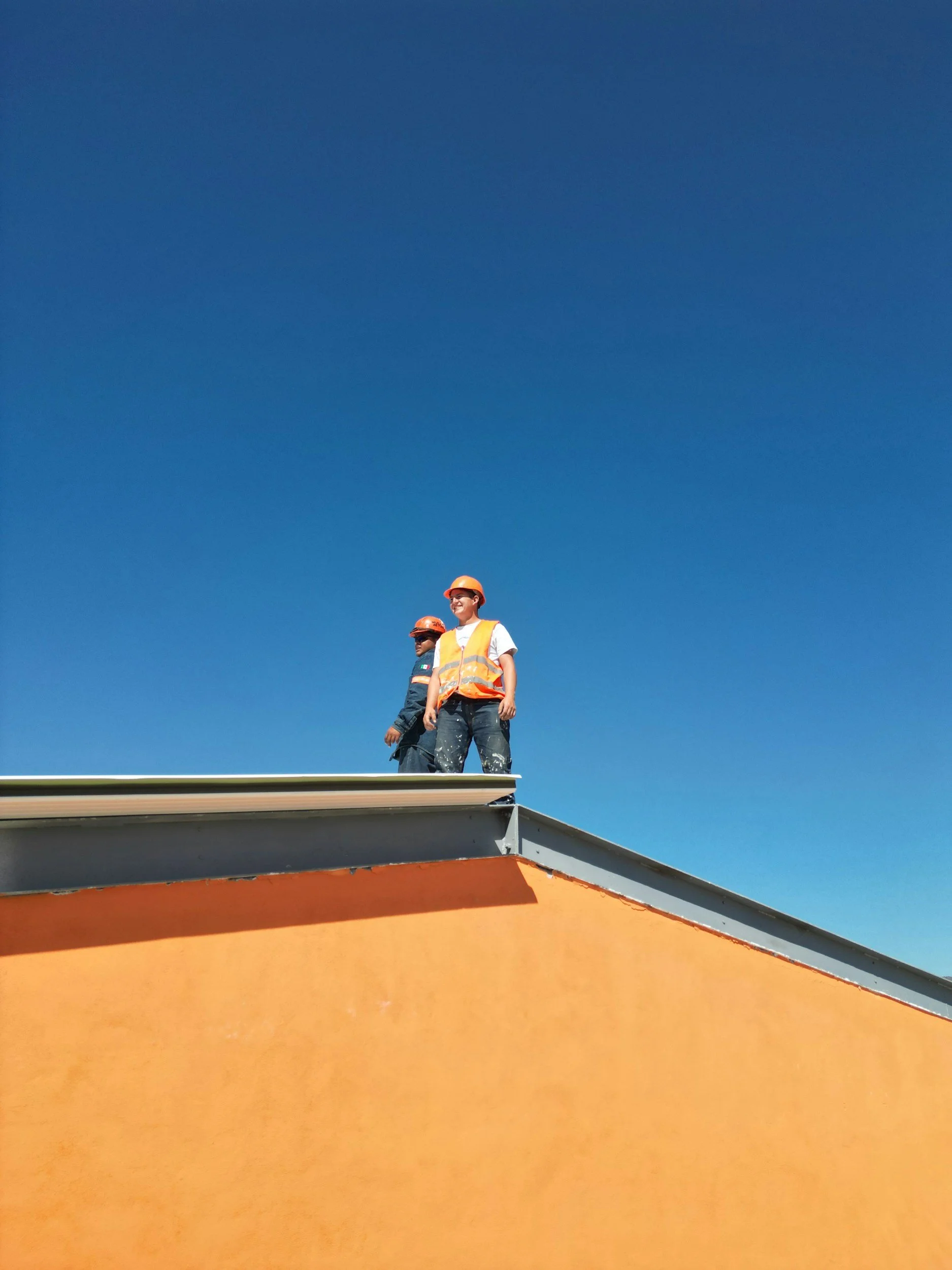 Two construction workers wearing orange safety vests and helmets standing on the roof of a building against a clear blue sky.