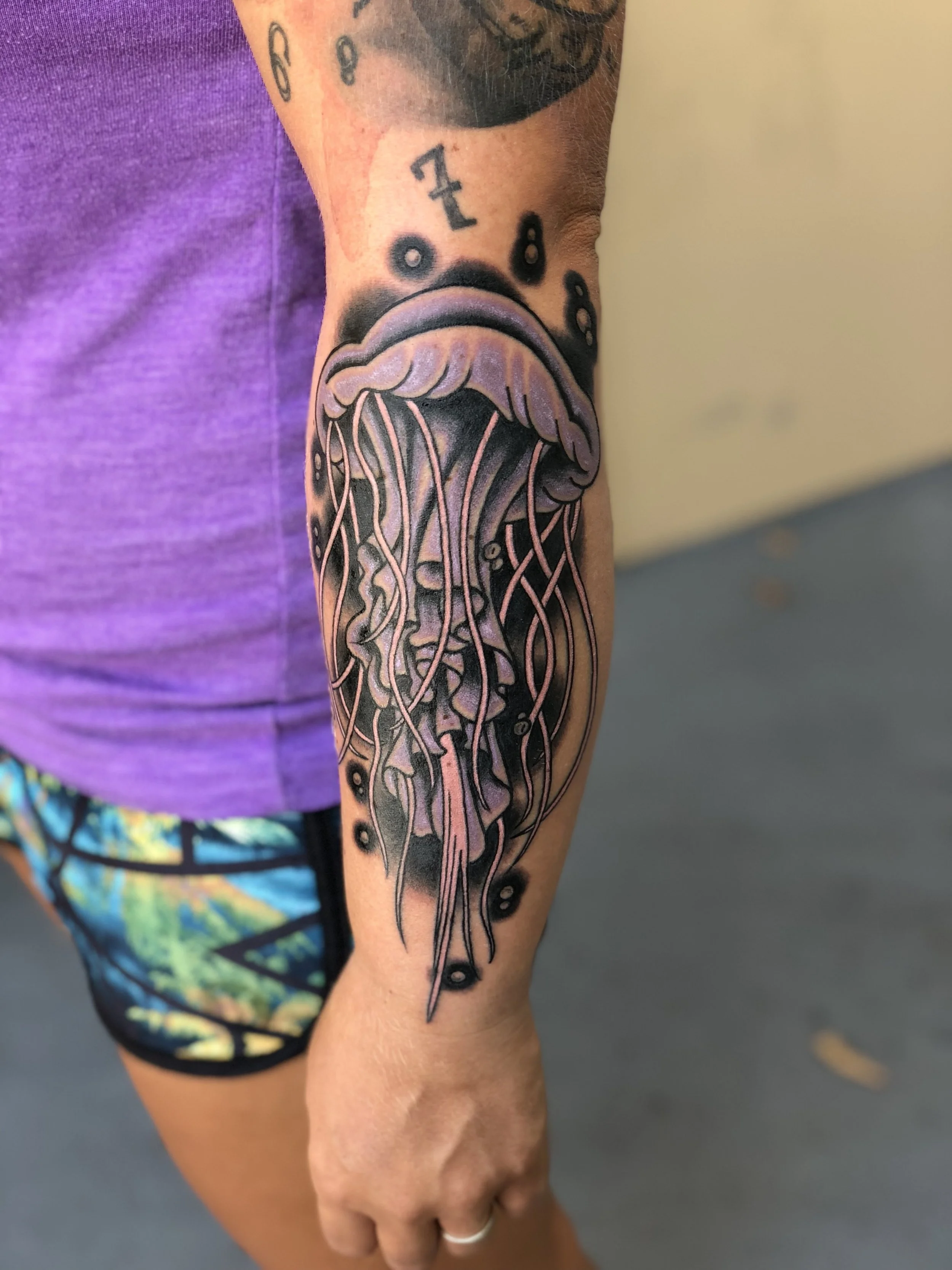 Jellyfish with trailing tentacles and soft rounded dome, rendered here in black light purple and deep black shading that gives it an almost glowing effect. Traditional American color forearm tattoo by Nick at Big Kahuna Tattoo in Boca Raton, FL.