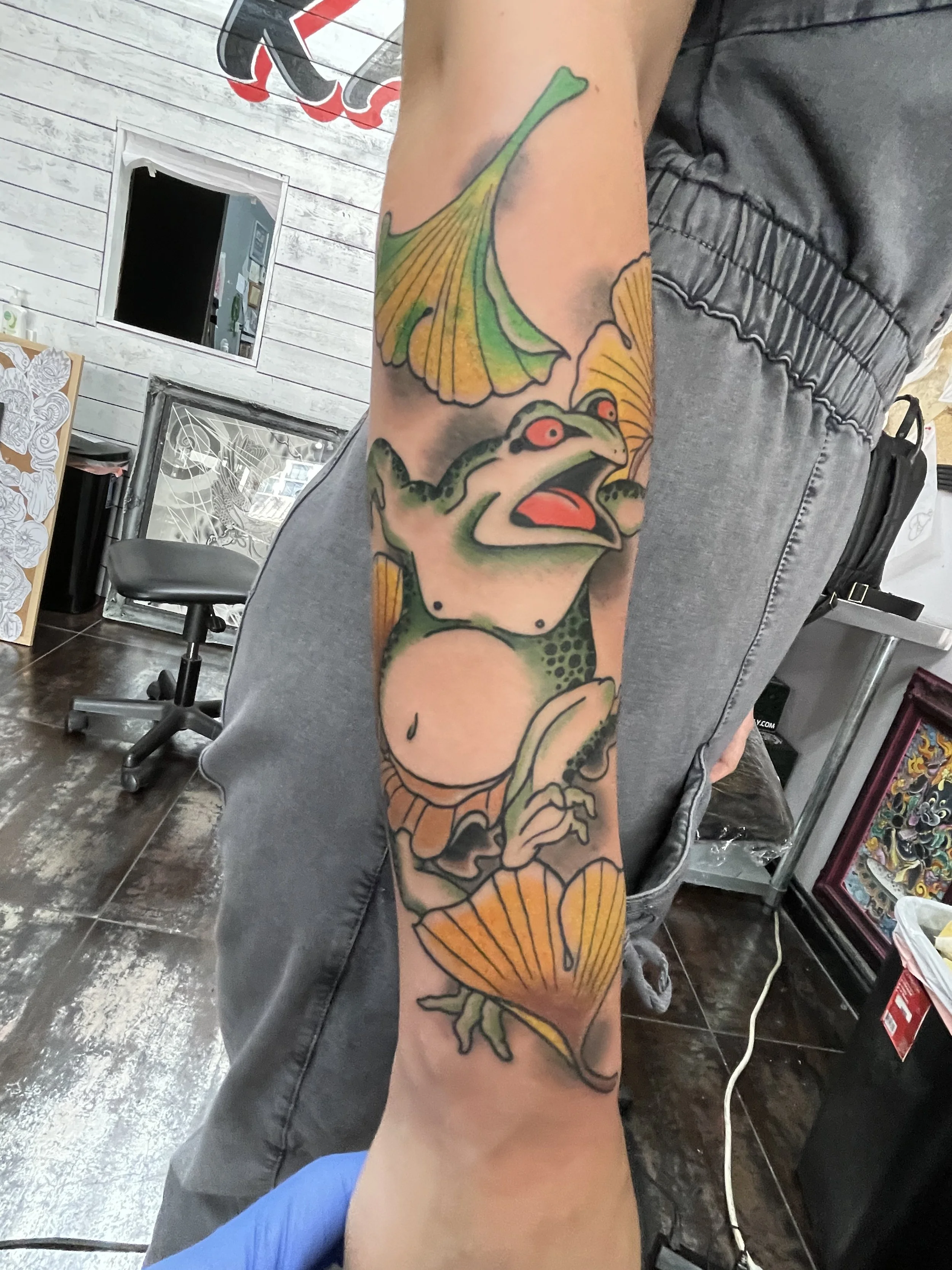 Japanese color forearm tattoo featuring a bold expressive frog with red eyes and an open mouth, surrounded by large fan-shaped golden ginkgo leaves cascading up and down the forearm, custom tattoo by Nick at Big Kahuna Tattoo in Boca Raton, FL.