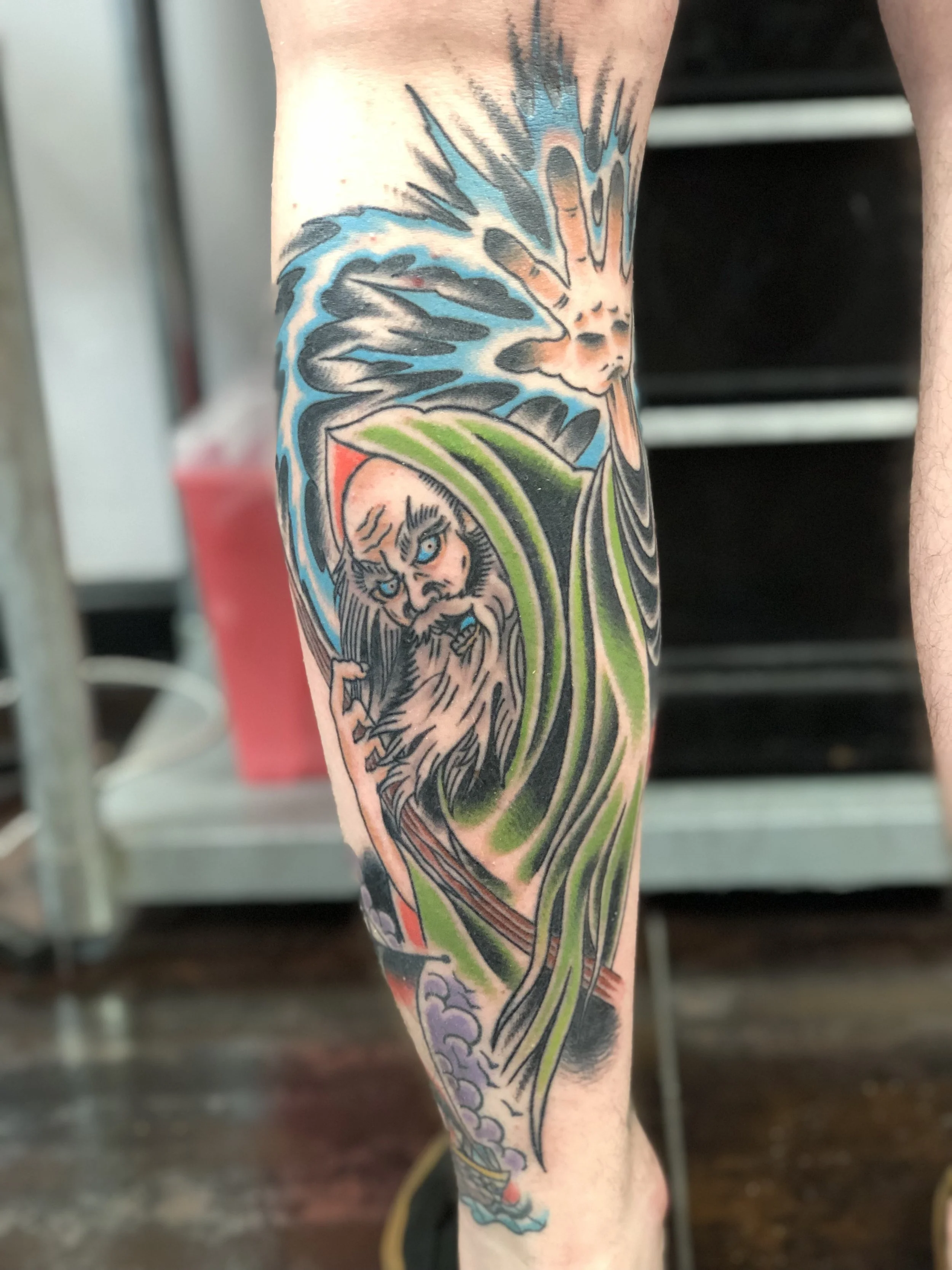 Traditional American color calf tattoo featuring a hooded wizard with piercing blue eyes, a long white beard, and a glowing sun or explosion of light behind him, custom tattoo by Nick at Big Kahuna Tattoo in Boca Raton, FL.