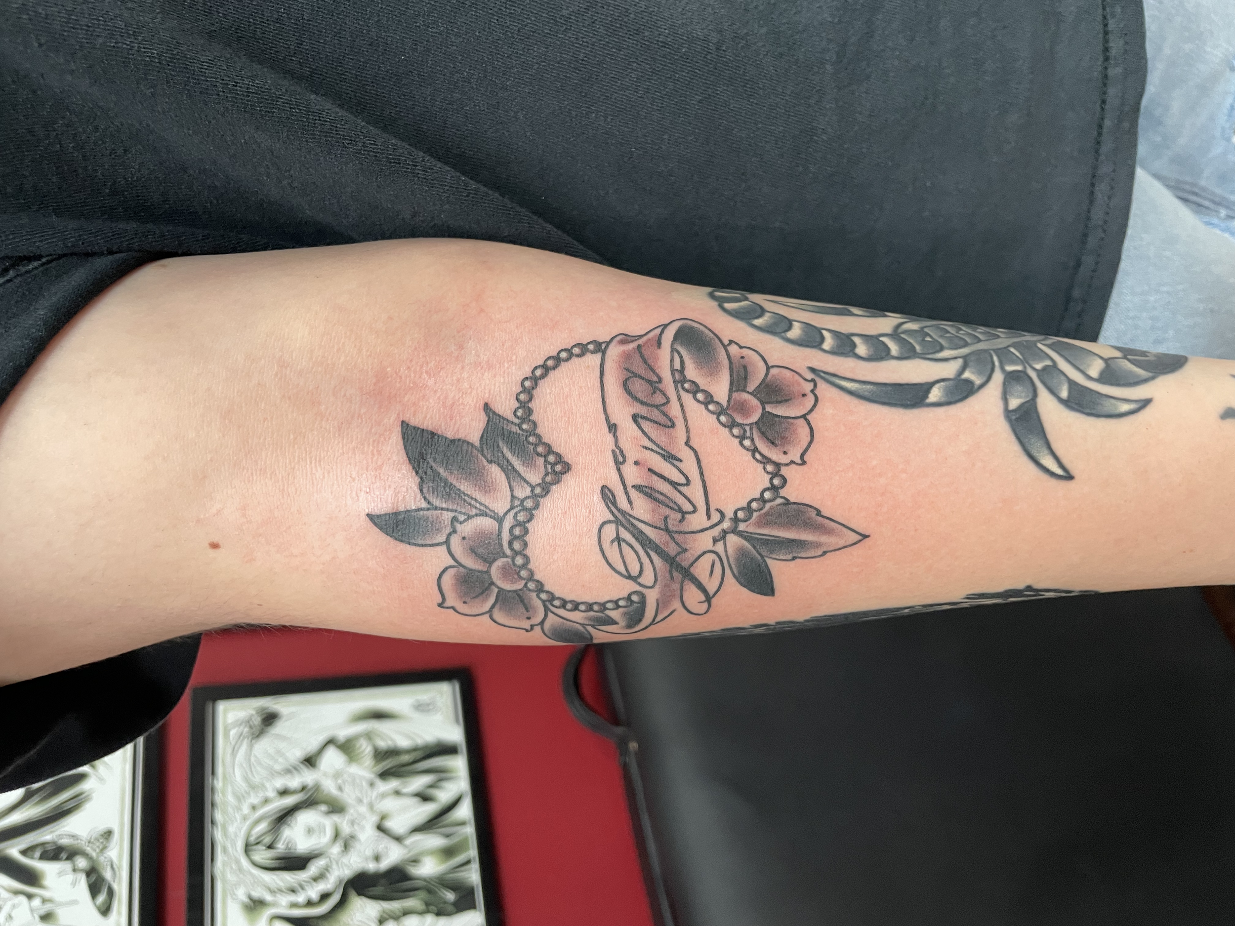 Traditional American black and grey forearm tattoo featuring a pearl beaded heart with flowers and leaves framing a banner script reading "Alina," custom tattoo by Nick at Big Kahuna Tattoo in Boca Raton, FL.