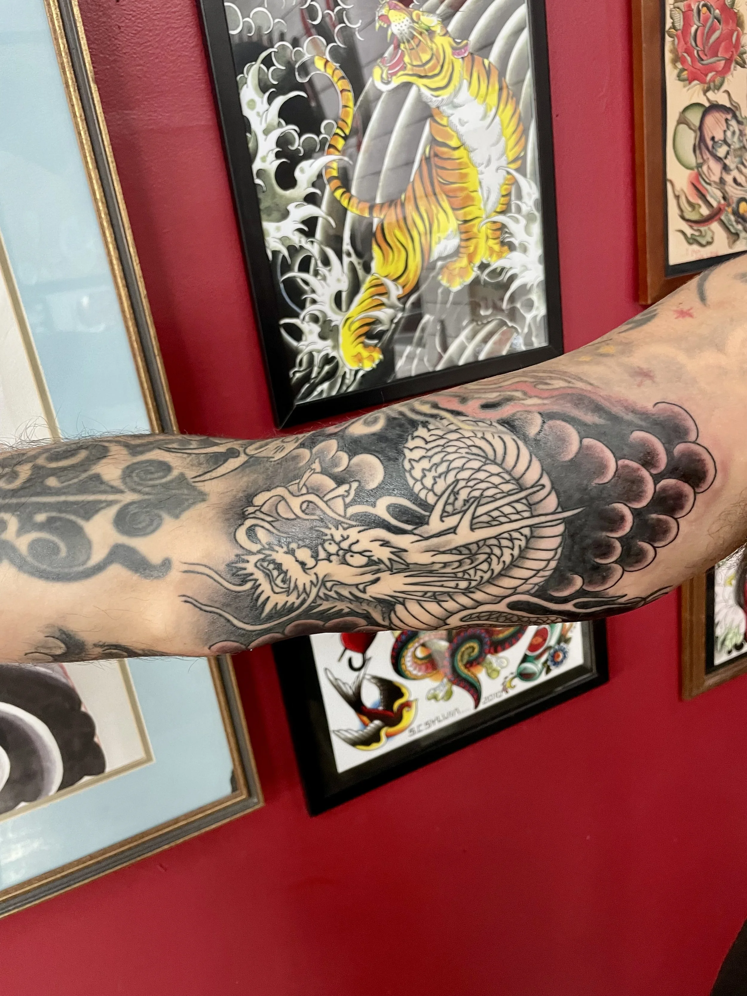 A pale dragon with detailed scales pushes through dark rolling clouds on the forearm, photographed against the shop's flash wall. Bold linework and strong shading that prove black and grey Japanese work has its own kind of power. Forearm tattoo by Ni