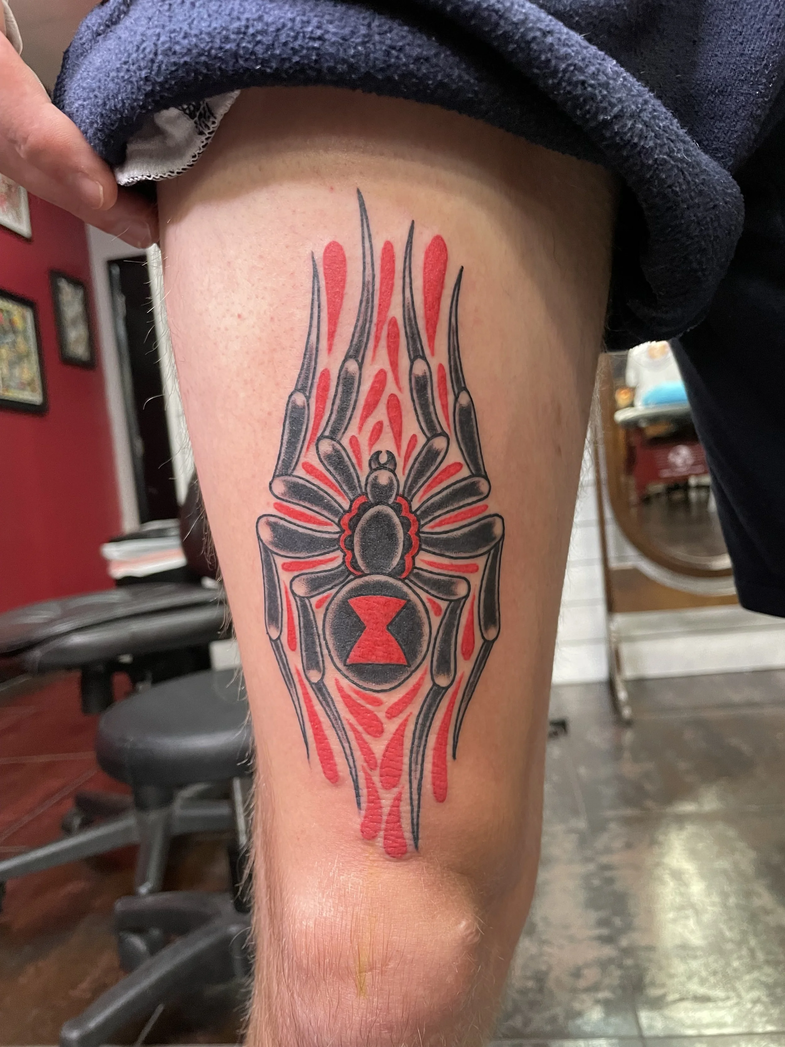 Bold traditional American thigh tattoo featuring a black widow spider with a red hourglass marking on its abdomen, surrounded by dramatic red blood drips radiating outward, custom tattoo by Nick at Big Kahuna Tattoo in Boca Raton, FL.