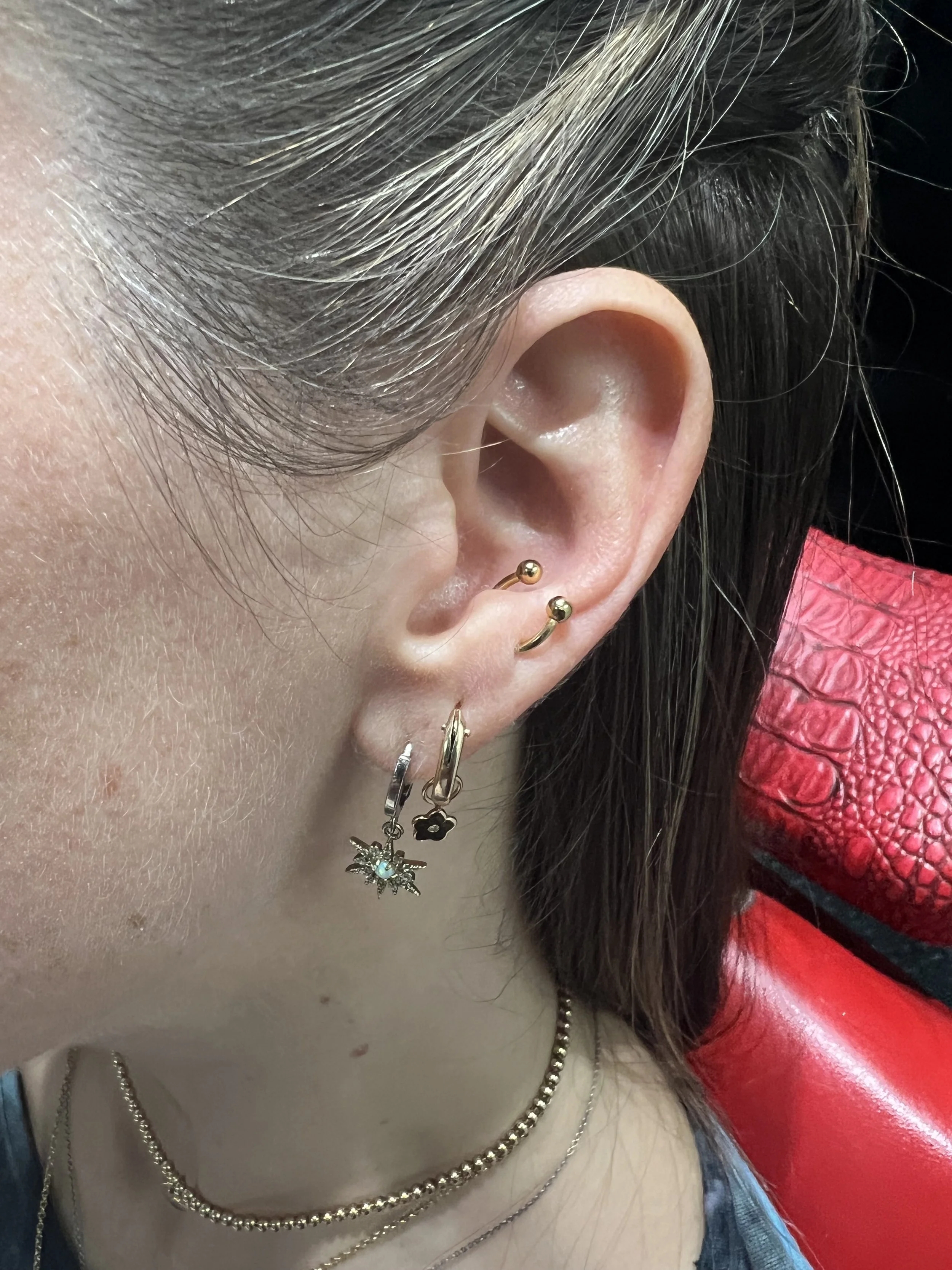 Tragus piercing with gold curved barbell, paired with two lobe piercings featuring charm hoops, a starburst with opal and a black flower dangle. Styled and pierced at Big Kahuna Tattoo in Boca Raton.