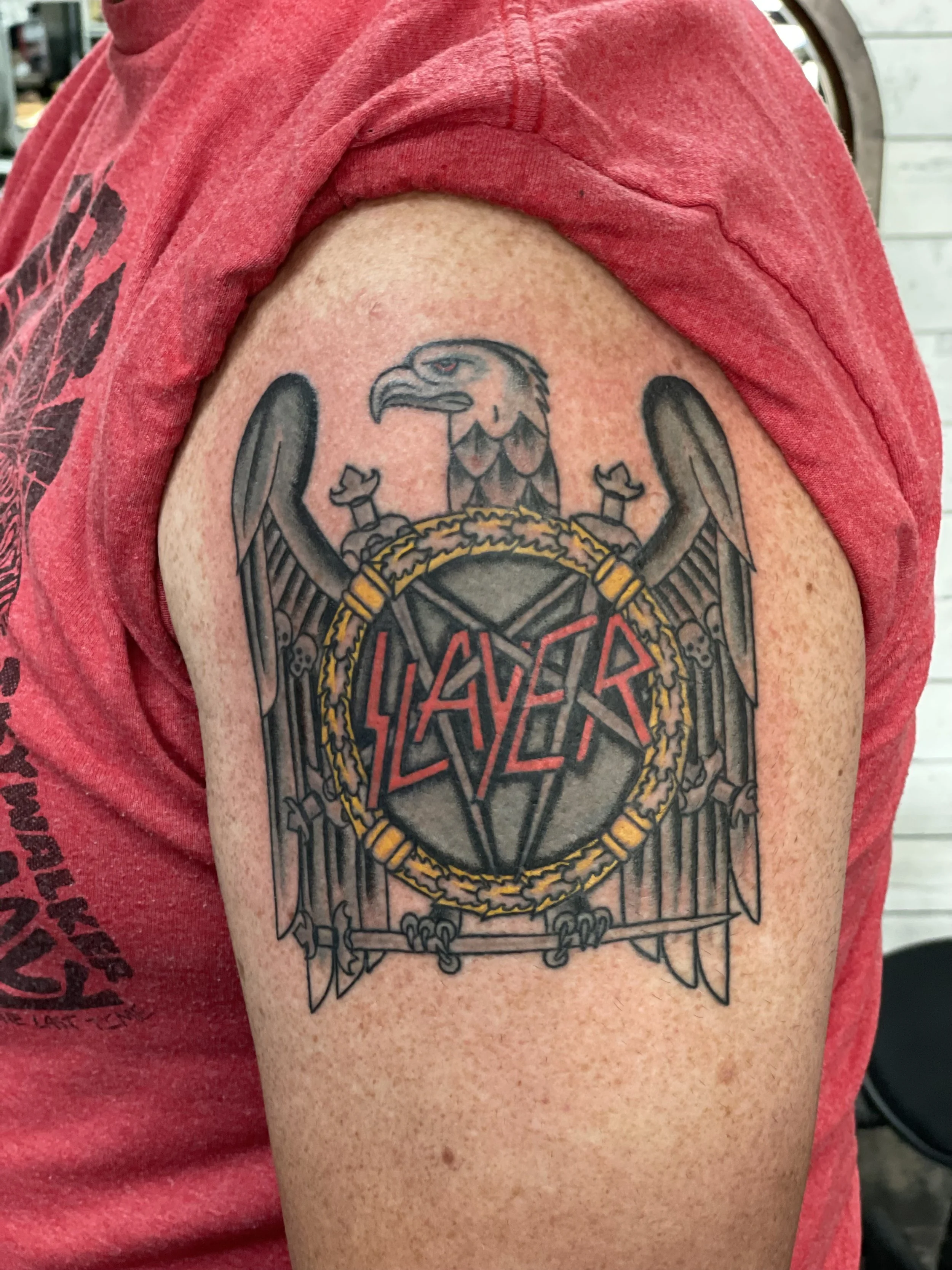 Color upper arm tattoo featuring the iconic Slayer band logo — a spread-winged eagle clutching swords over a pentagram shield ringed in gold laurels with the Slayer name in bold red lettering, custom tattoo by Nick at Big Kahuna Tattoo in Boca Raton,