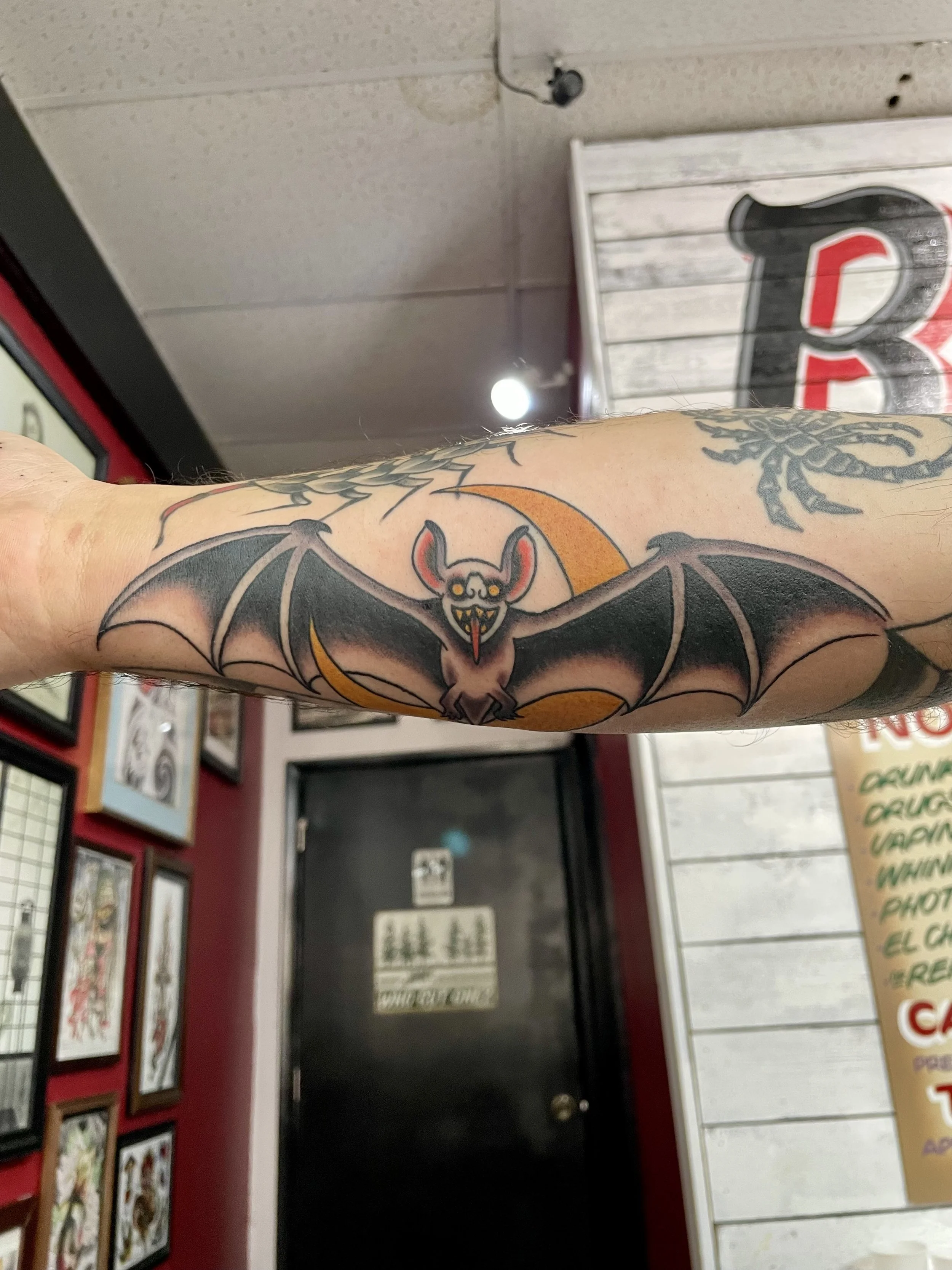 Wings stretched wide across the forearm, this traditional bat with fanged grin and red ears hangs in front of a glowing gold moon. A Halloween staple executed with the kind of clean linework that makes it look good year round. Color forearm tattoo by