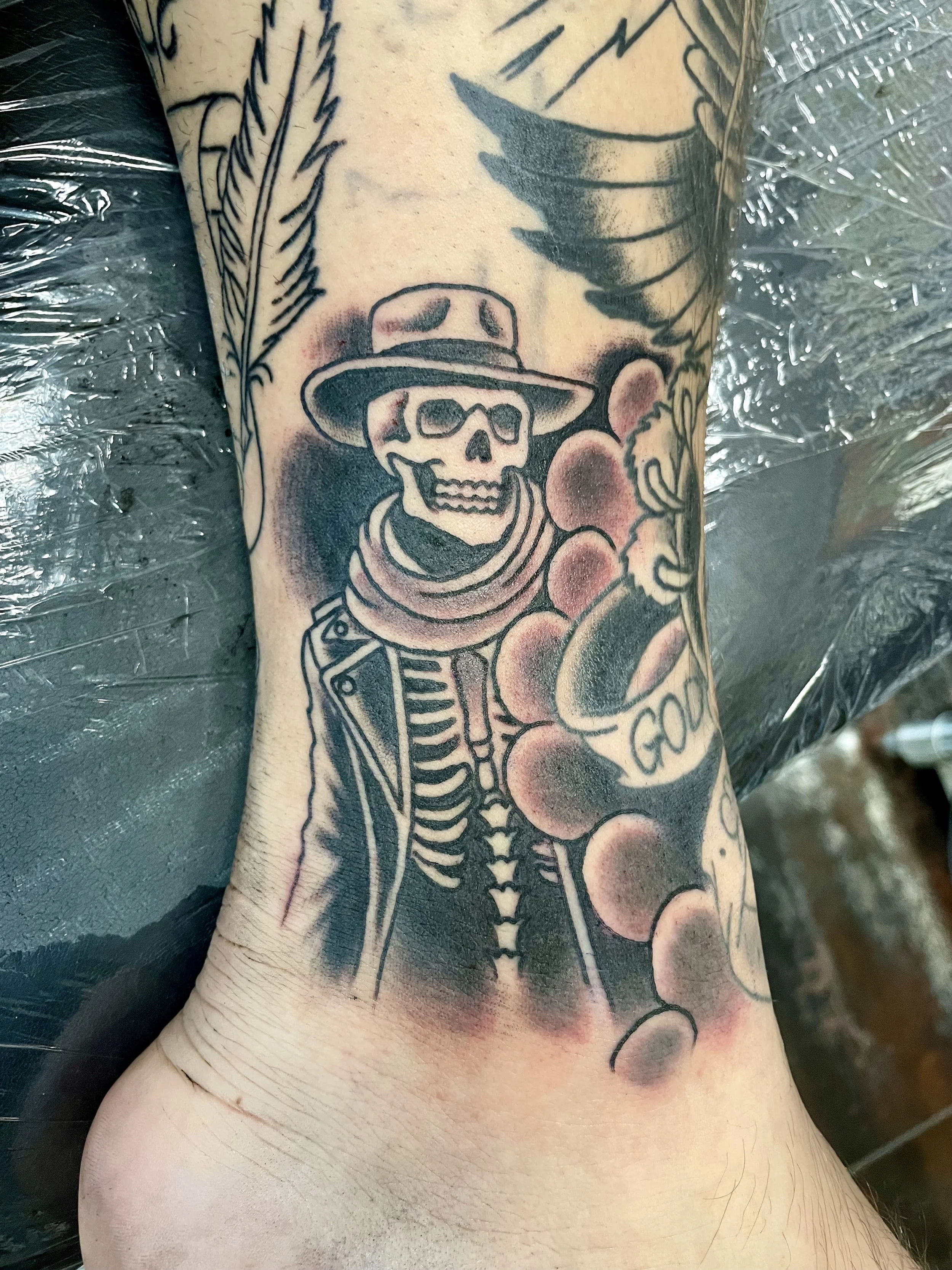 Black and grey ankle tattoo featuring a skeleton dressed in a cowboy hat and duster coat, set against a smoky dark background with surrounding sleeve work, custom tattoo by Nick at Big Kahuna Tattoo in Boca Raton, FL.