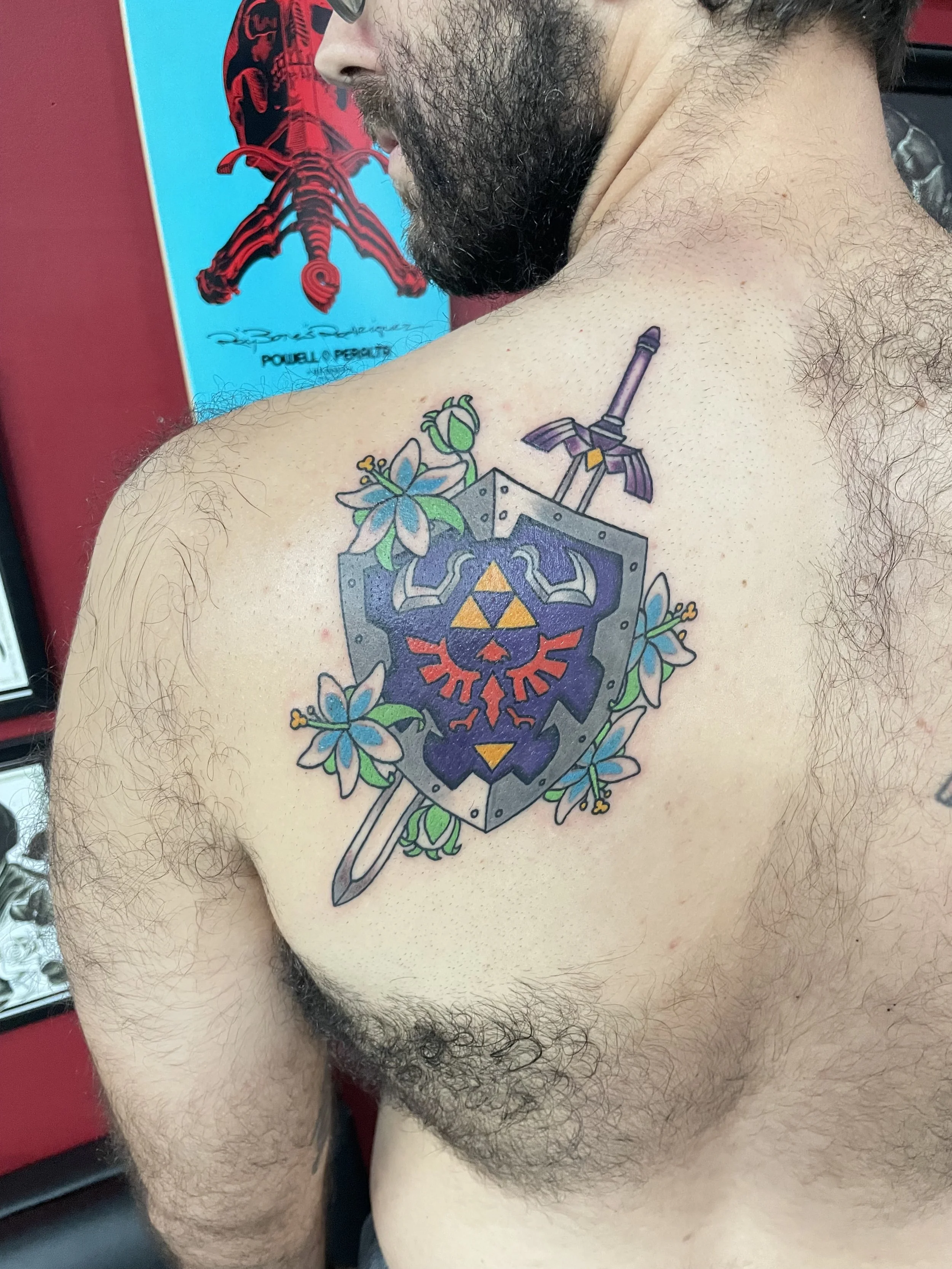 
Color back tattoo featuring the Legend of Zelda Hylian Shield with the Triforce and Royal Crest in blue, gold, and red, crossed with the Master Sword and surrounded by delicate blue Navi-inspired flowers, custom tattoo by Nick at Big Kahuna Tattoo i