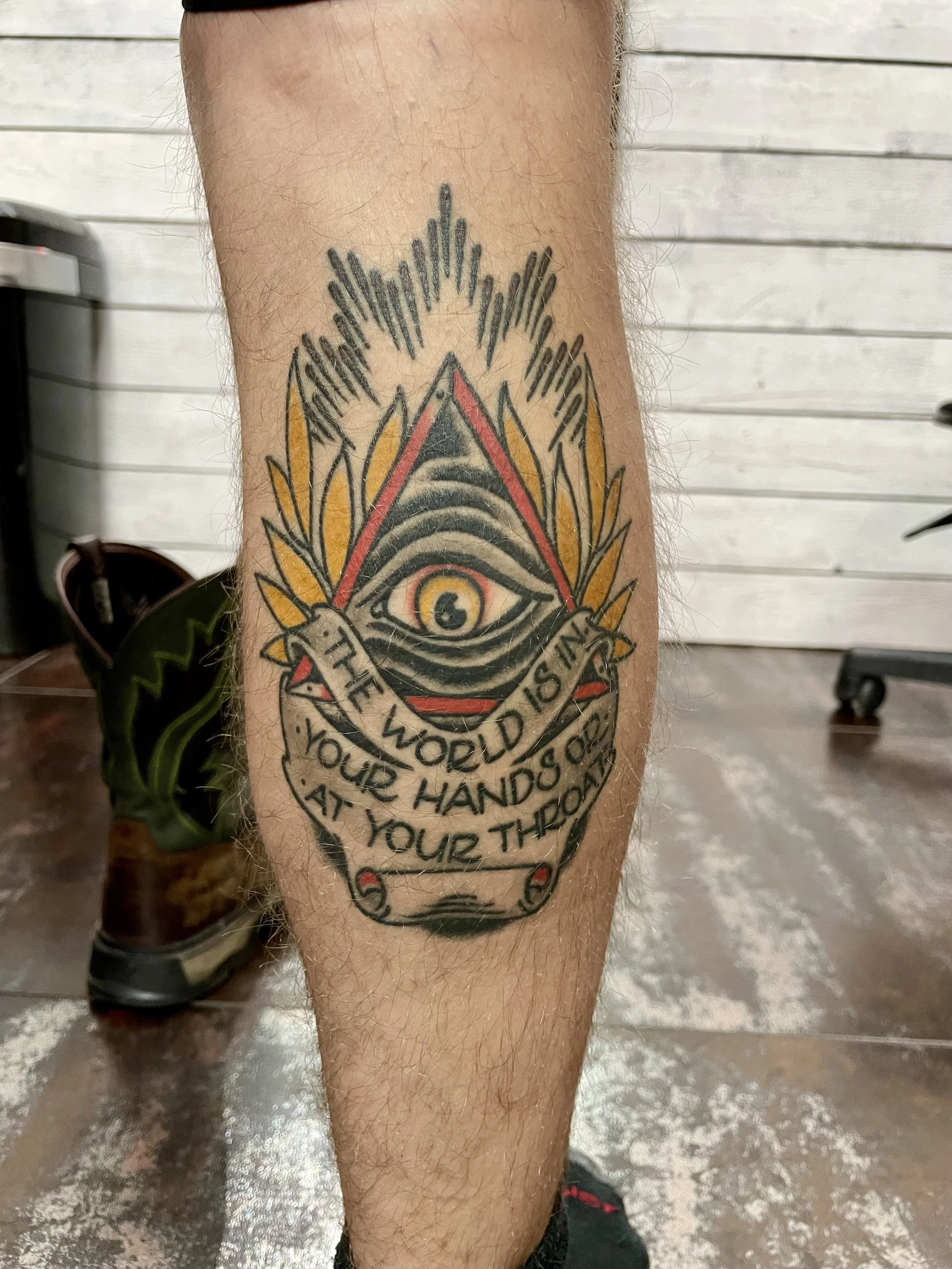 Traditional American color leg tattoo featuring an all-seeing eye in a red-bordered pyramid surrounded by gold leaves and radiating lines, with a scroll banner below reading "The World Is In Your Hands Or At Your Throat," custom tattoo by Nick at Big
