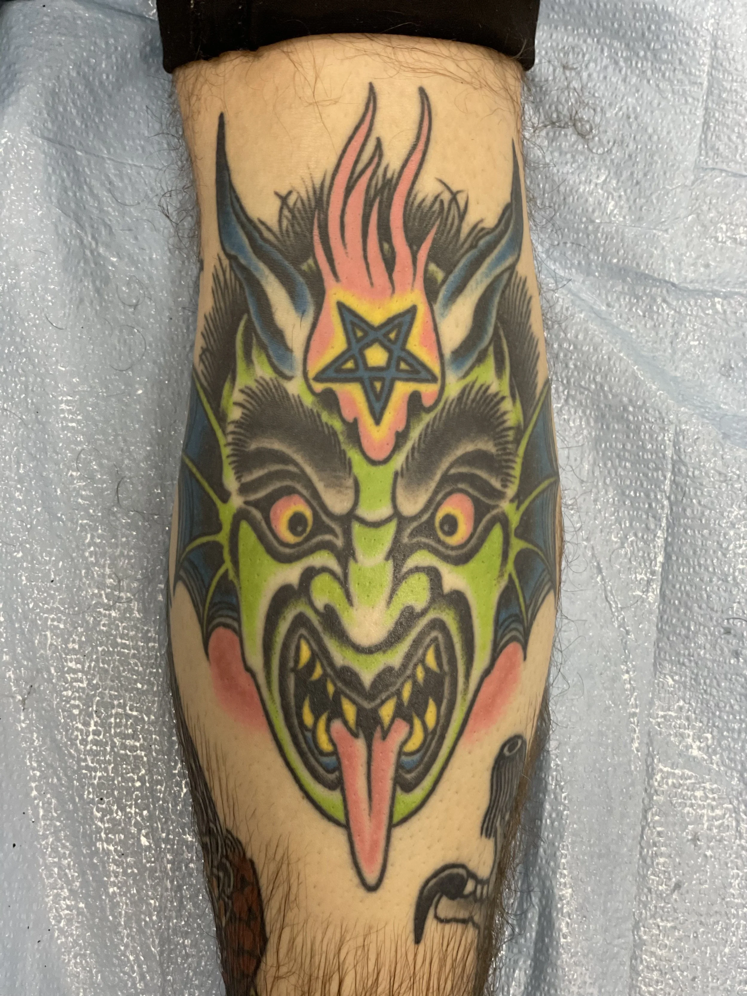 Bold traditional American color shin tattoo featuring a large green devil face with red eyes and a forked tongue, crowned with a blue pentagram star and pink flames rising from the top, with blue bat wings flanking the head, custom tattoo by Nick at 