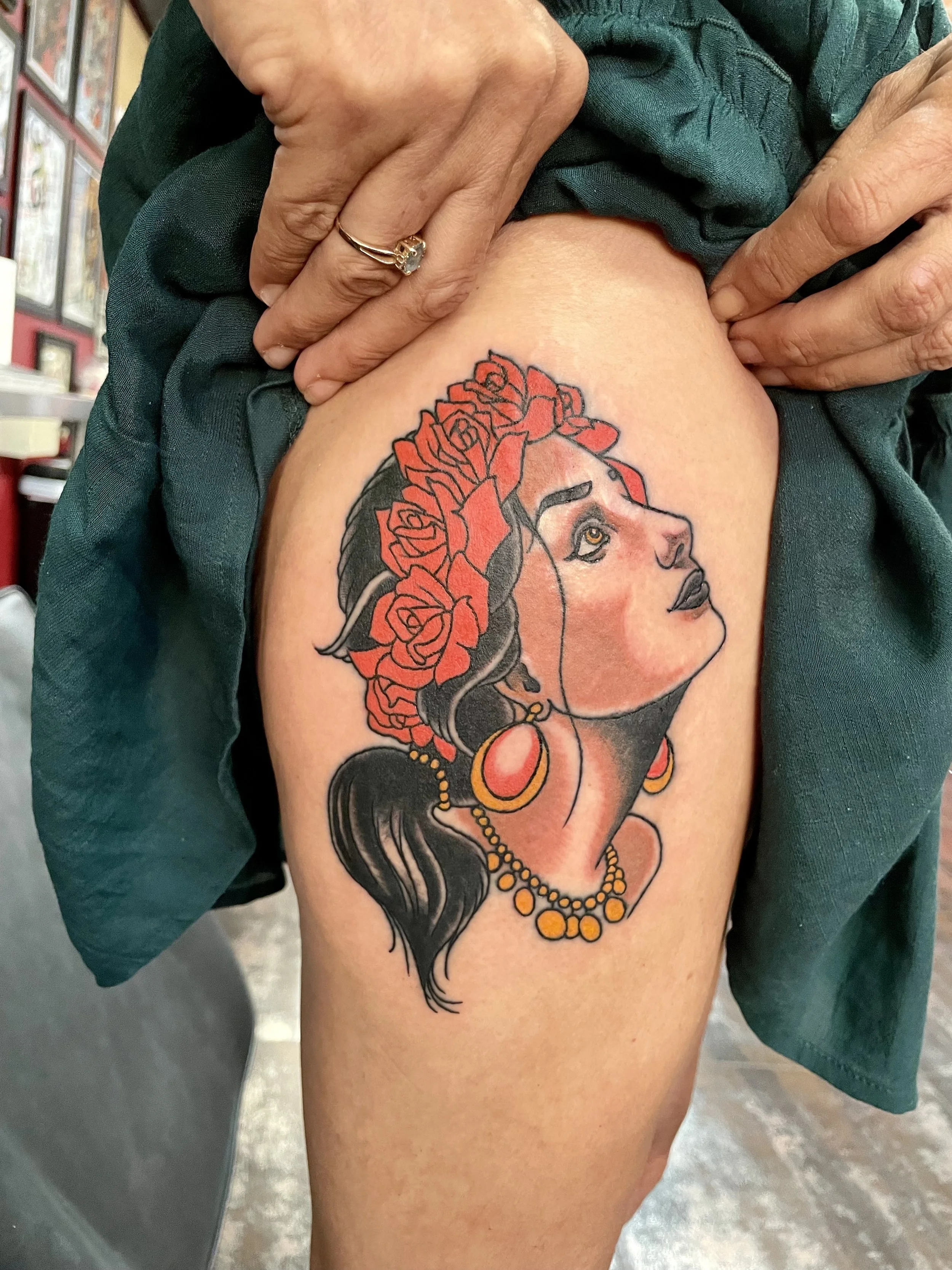 Traditional American color thigh tattoo featuring a classic gypsy woman portrait in profile with dark hair, golden hoop earrings, a beaded necklace, and a crown of bold red roses, custom tattoo by Nick at Big Kahuna Tattoo in Boca Raton, FL.