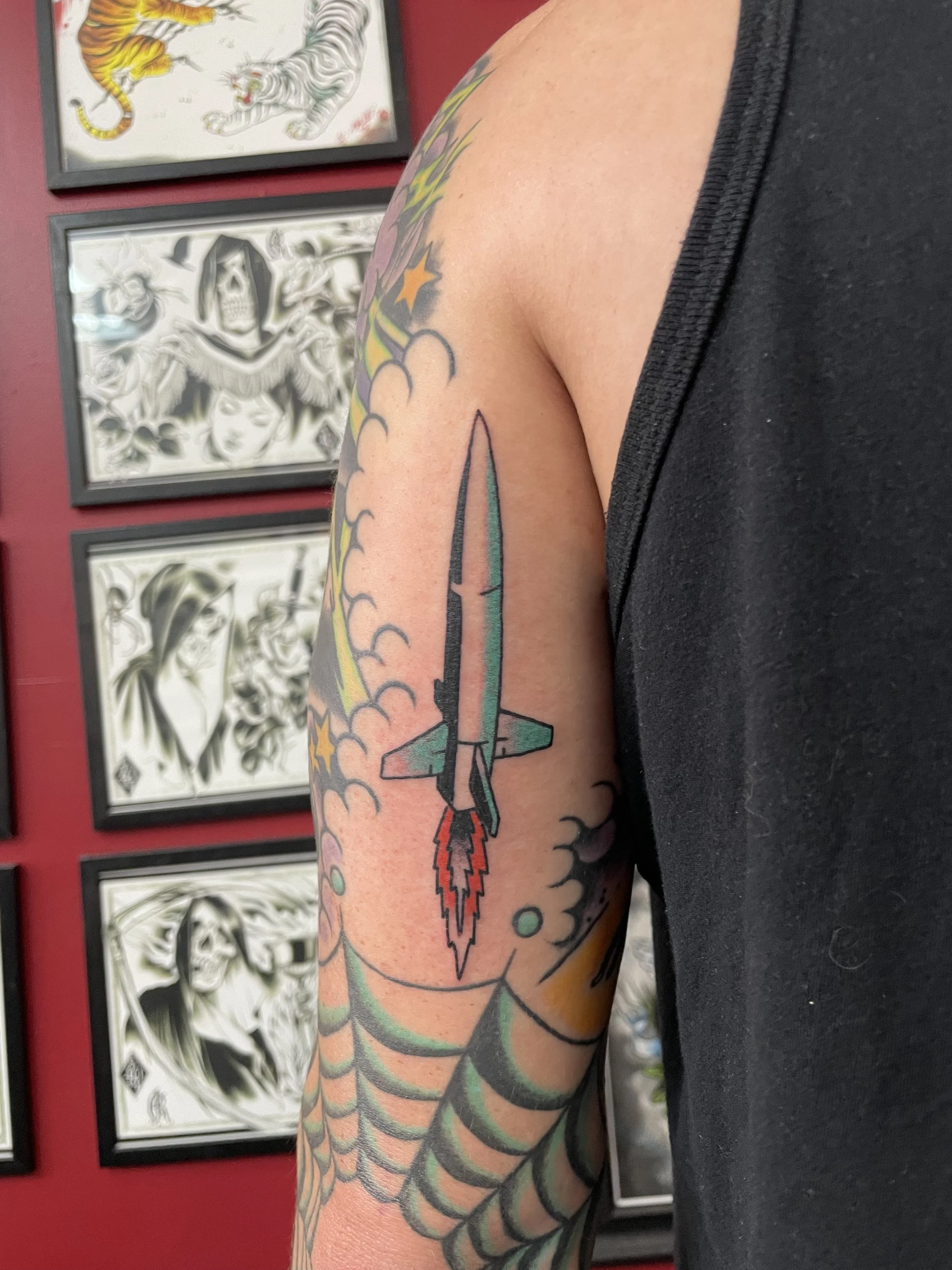 Traditional American color sleeve tattoo featuring a sleek rocket ship launching upward through billowing clouds with a red flame exhaust, set among existing sleeve work including gold stars and a spiderweb, custom tattoo by Nick at Big Kahuna Tattoo