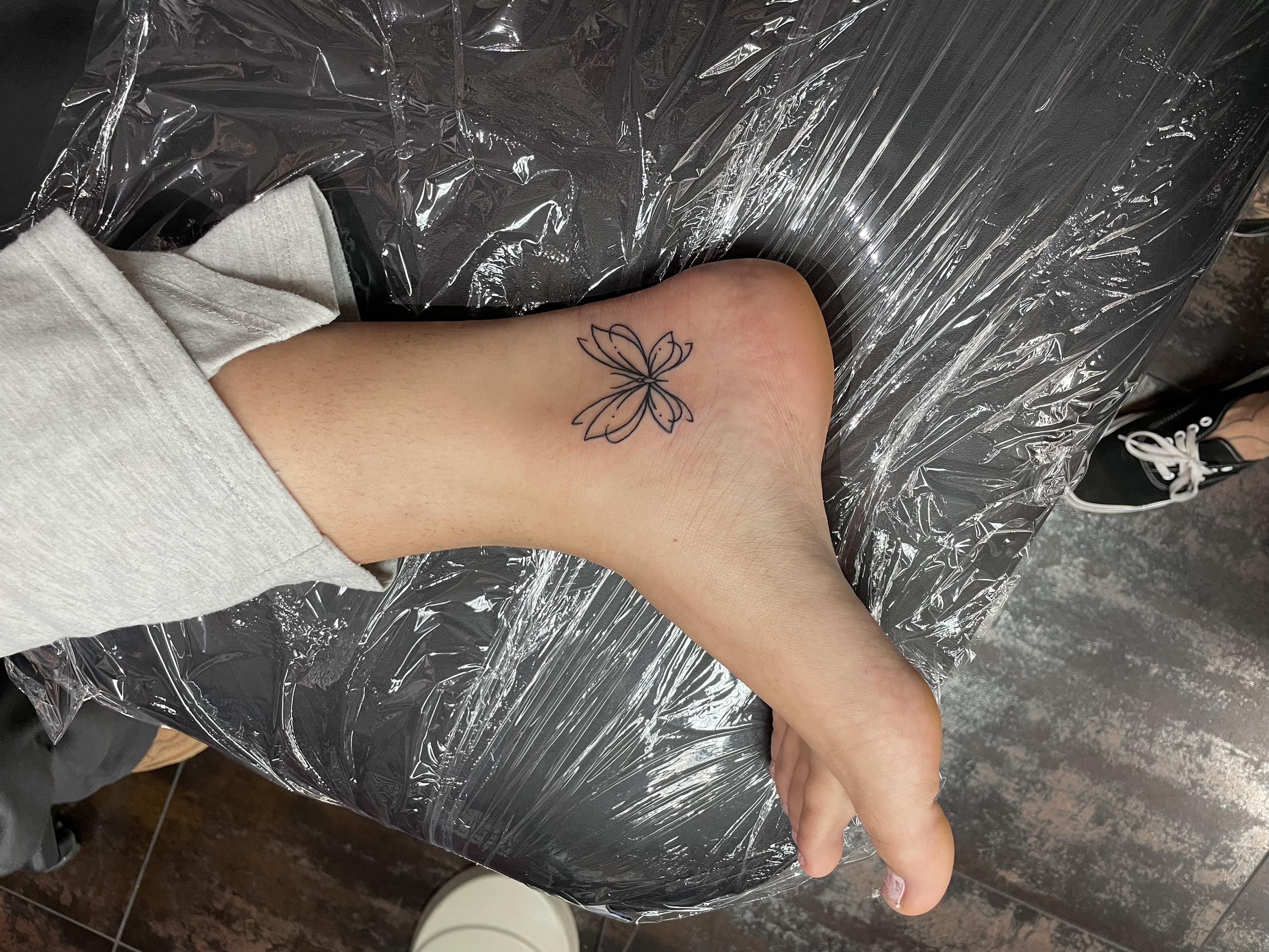 Delicate fine line ankle tattoo featuring a small minimalist butterfly outline in a clean single stroke style, custom tattoo by Nick at Big Kahuna Tattoo in Boca Raton, FL.