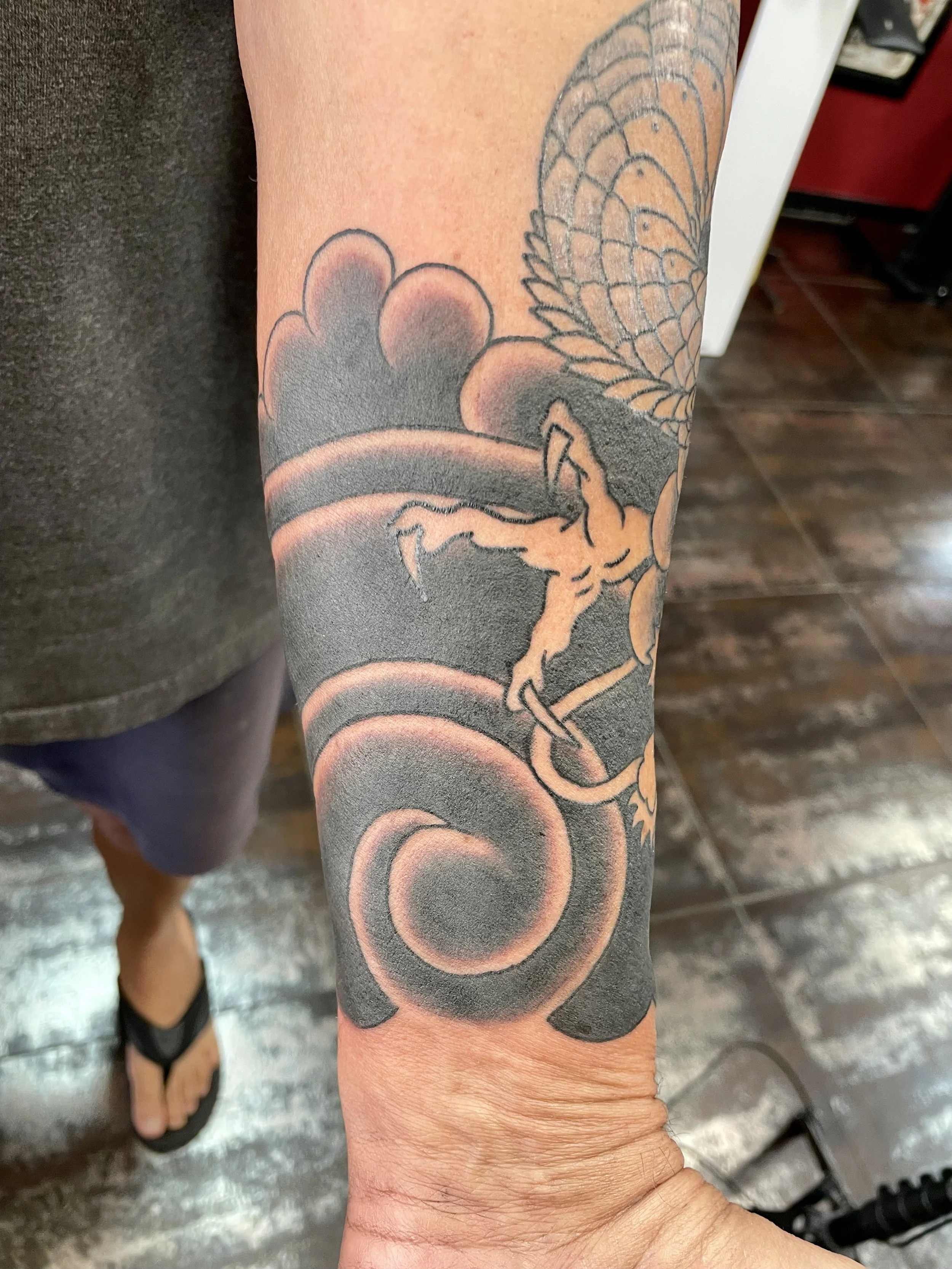 Japanese black and grey forearm tattoo featuring a coiling dragon emerging from swirling wind clouds, with richly detailed scales and a fierce expression, custom tattoo by Nick at Big Kahuna Tattoo in Boca Raton, FL.
