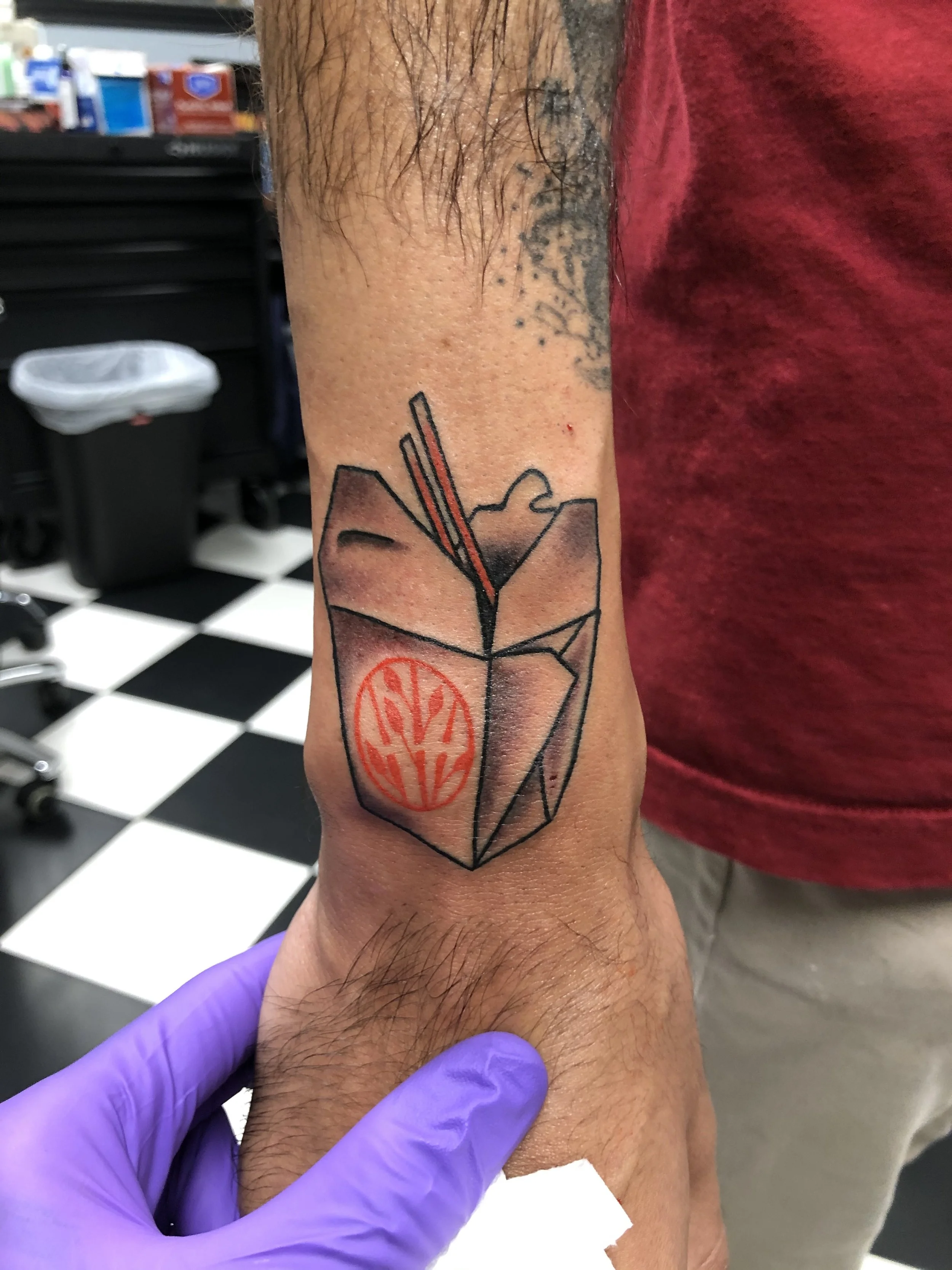 Minimalist black and red wrist tattoo featuring a Chinese takeout box with chopsticks and a red stamp logo, custom tattoo by Nick at Big Kahuna Tattoo in Boca Raton, FL.