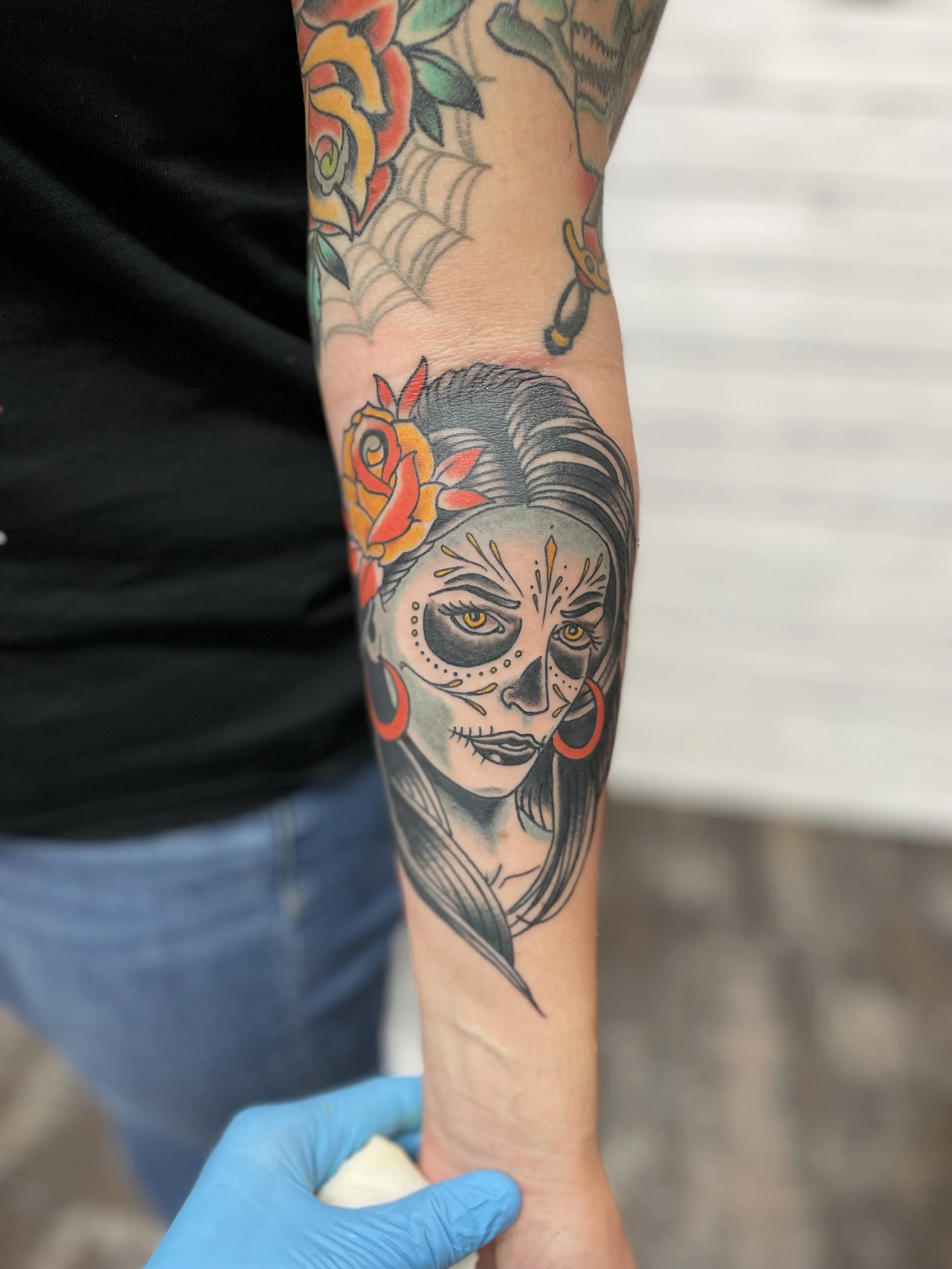 Traditional American forearm tattoo featuring a sugar skull woman with golden eyes, intricate face paint markings, red hoop earrings, and a vibrant orange and red rose tucked in her dark hair, rendered in a striking combination of black, grey, and bo