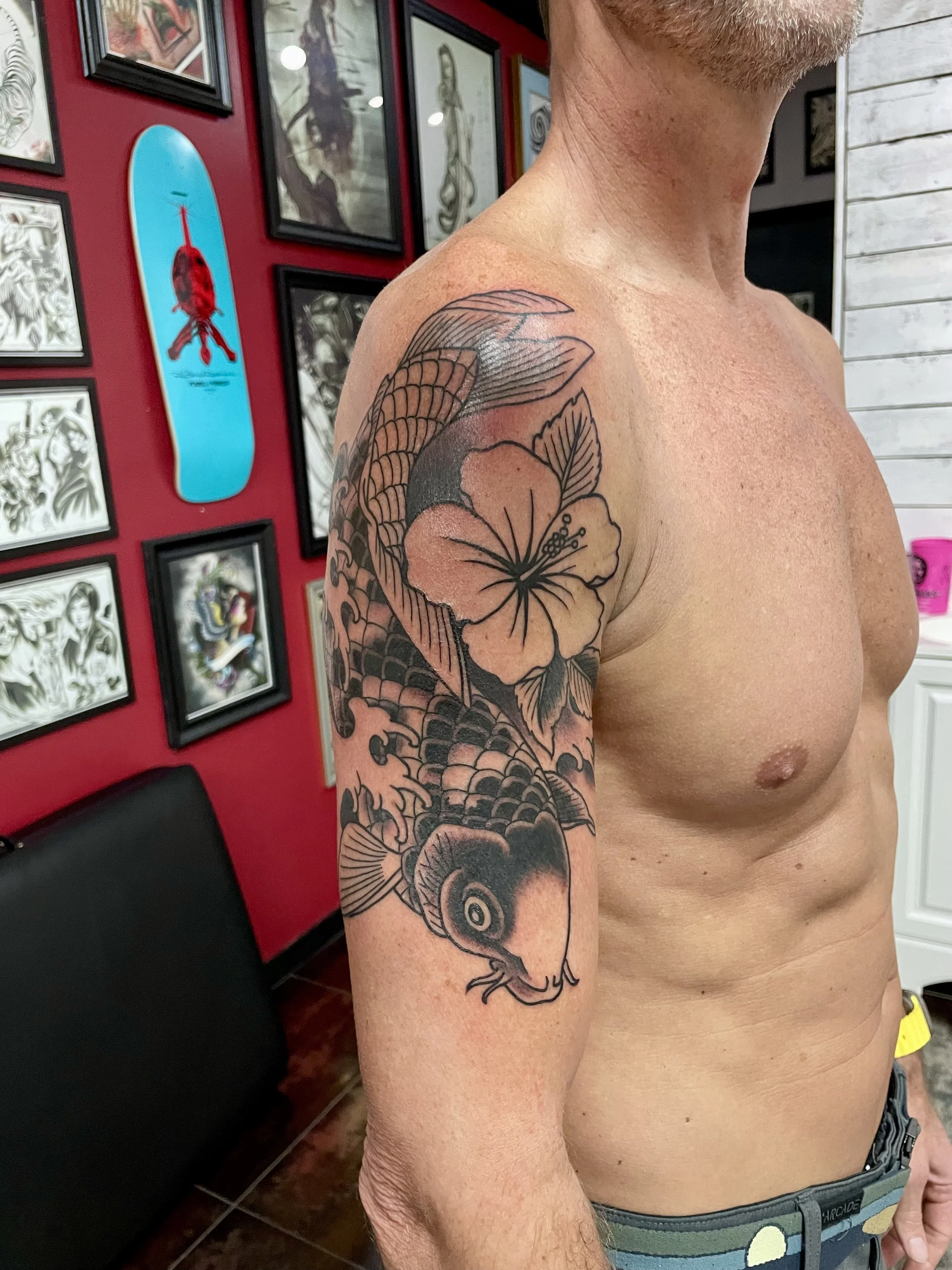 Japanese black and grey upper arm tattoo featuring a large koi fish with detailed scales curving around the shoulder, accompanied by a bold hibiscus flower and flowing water elements, custom tattoo by Nick at Big Kahuna Tattoo in Boca Raton, FL.