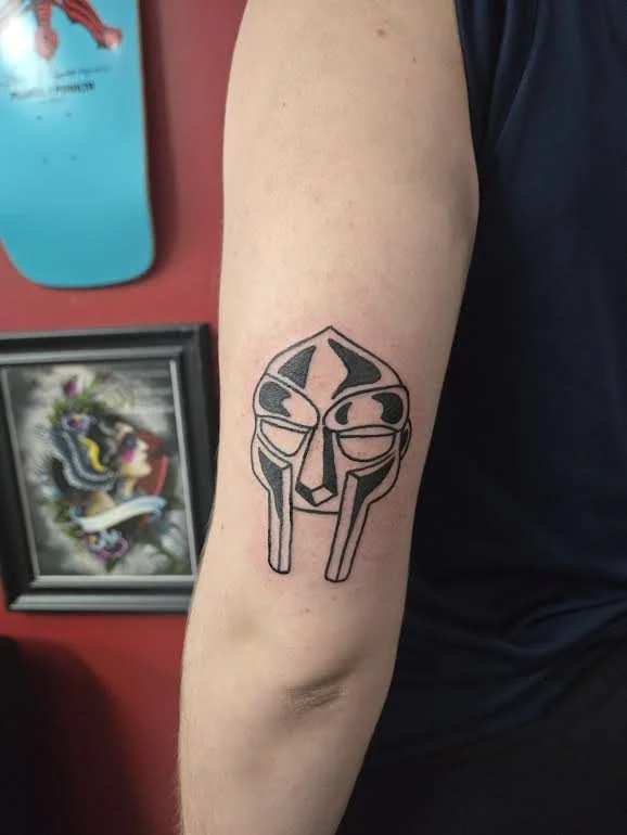 Black and grey traditional upper arm tattoo featuring the iconic MF DOOM metal mask, custom music tribute tattoo by Brandon Perez at Big Kahuna Tattoo in Boca Raton, FL