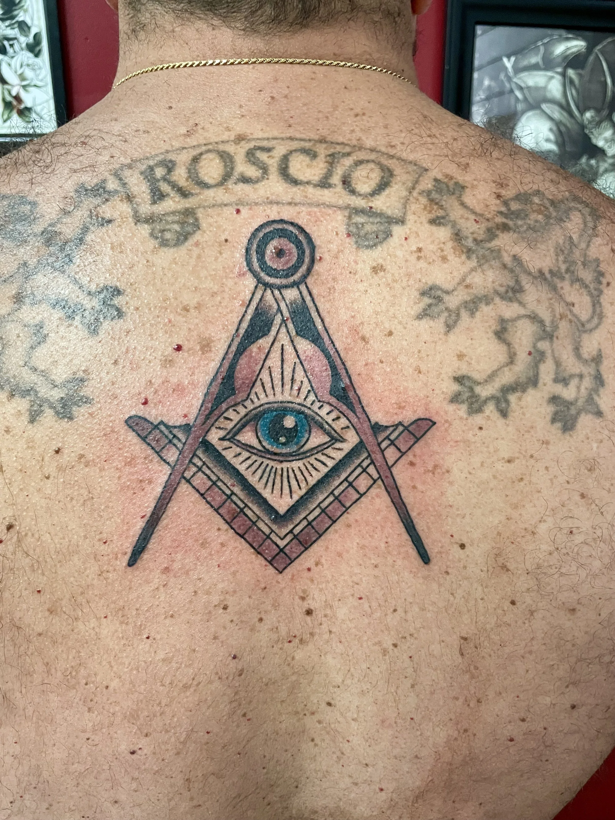 Black and grey upper back tattoo featuring the Freemason square and compass symbol with a detailed all-seeing eye in a pyramid at the center, rendered with bold linework and subtle color shading, custom tattoo by Nick at Big Kahuna Tattoo in Boca Rat