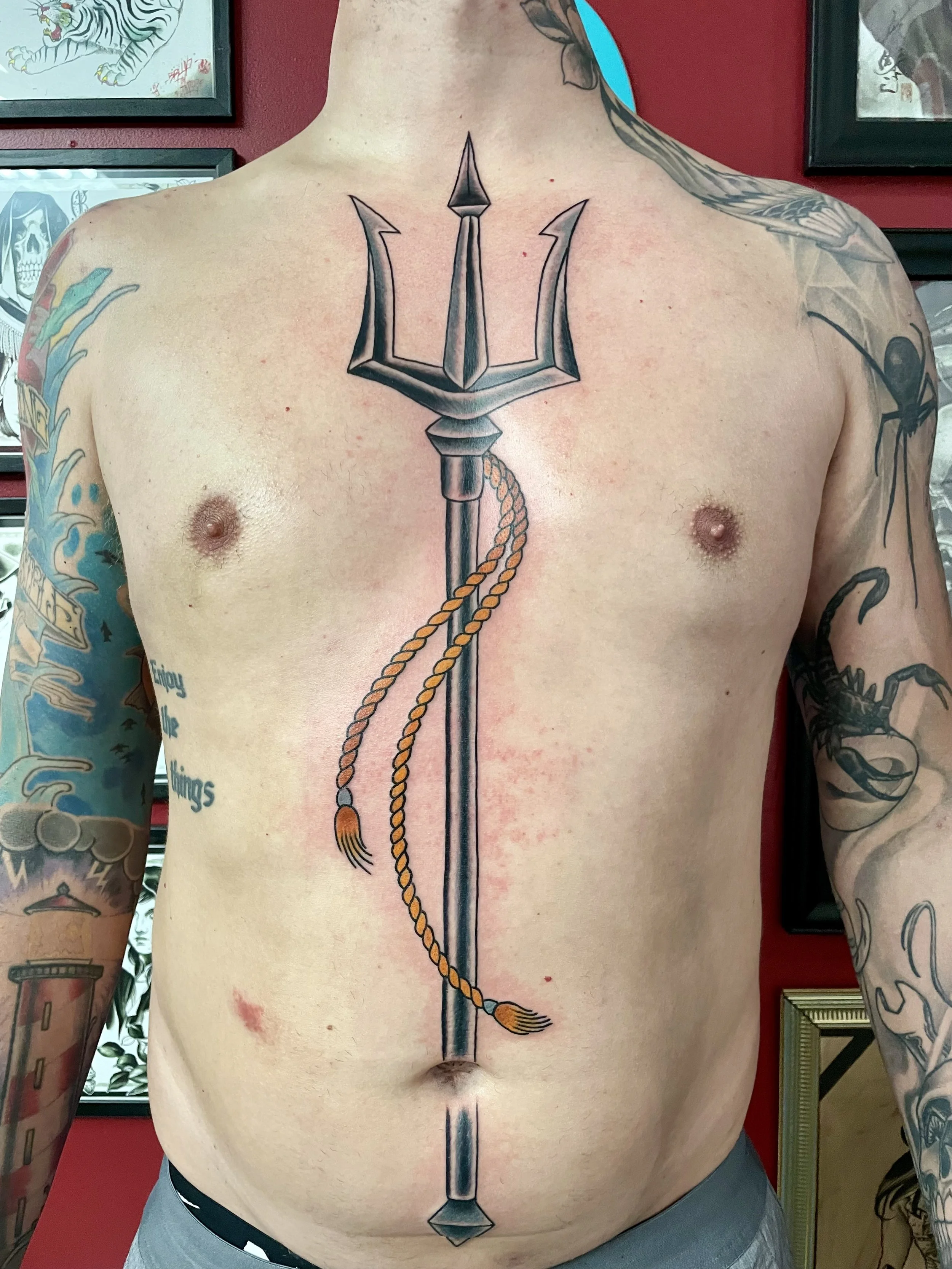 Large black and grey sternum and stomach tattoo featuring Poseidon's trident running vertically from chest to below the navel, with a detailed three-pronged tip and a braided gold rope coiled around the shaft with tasseled ends, custom tattoo by Nick