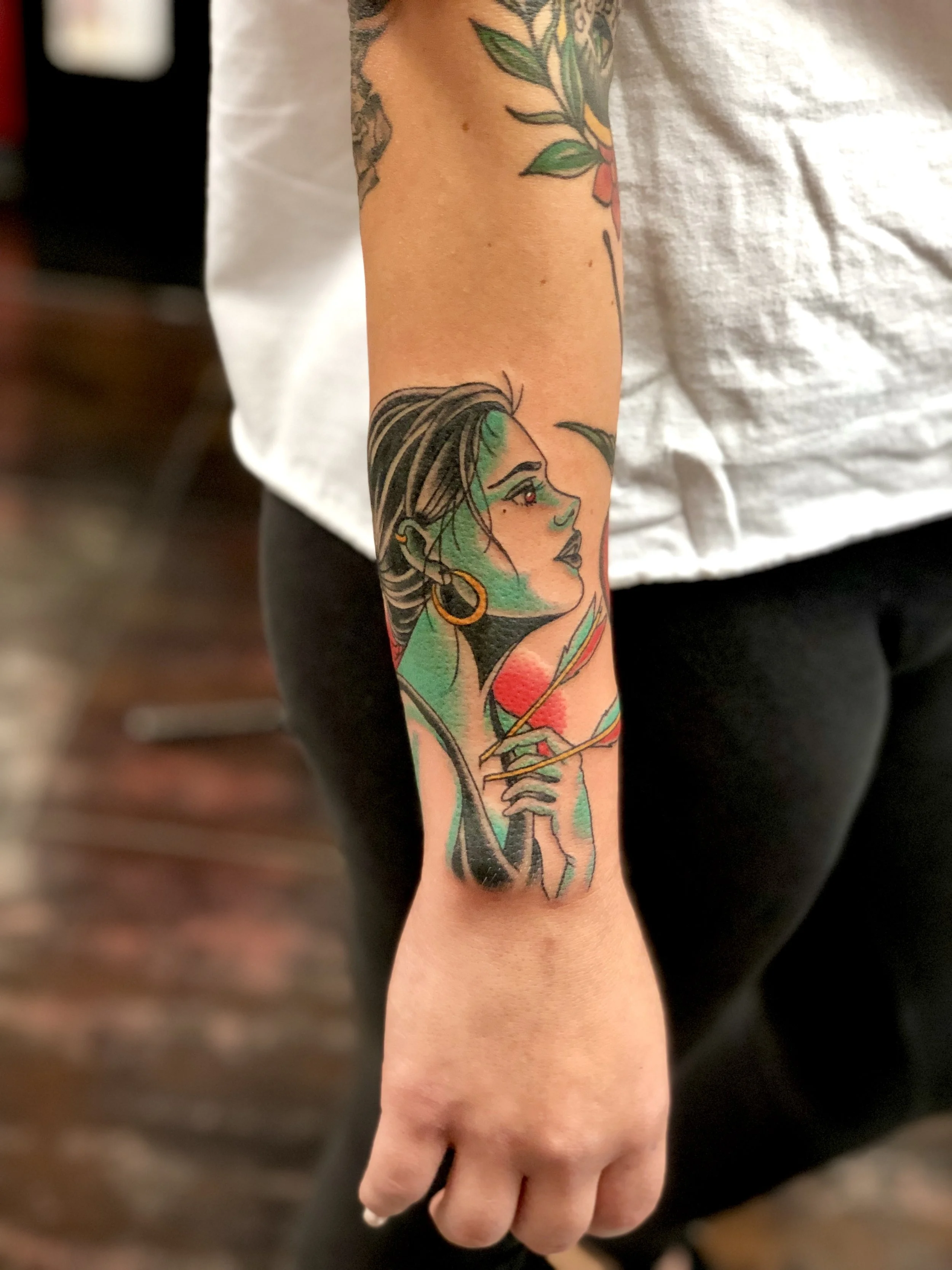 Traditional American color tattoo featuring a green-skinned gypsy woman in side profile holding a feather, with gold hoop earring and bold linework, tattooed on the forearm by Nick at Big Kahuna Tattoo in Boca Raton, FL.