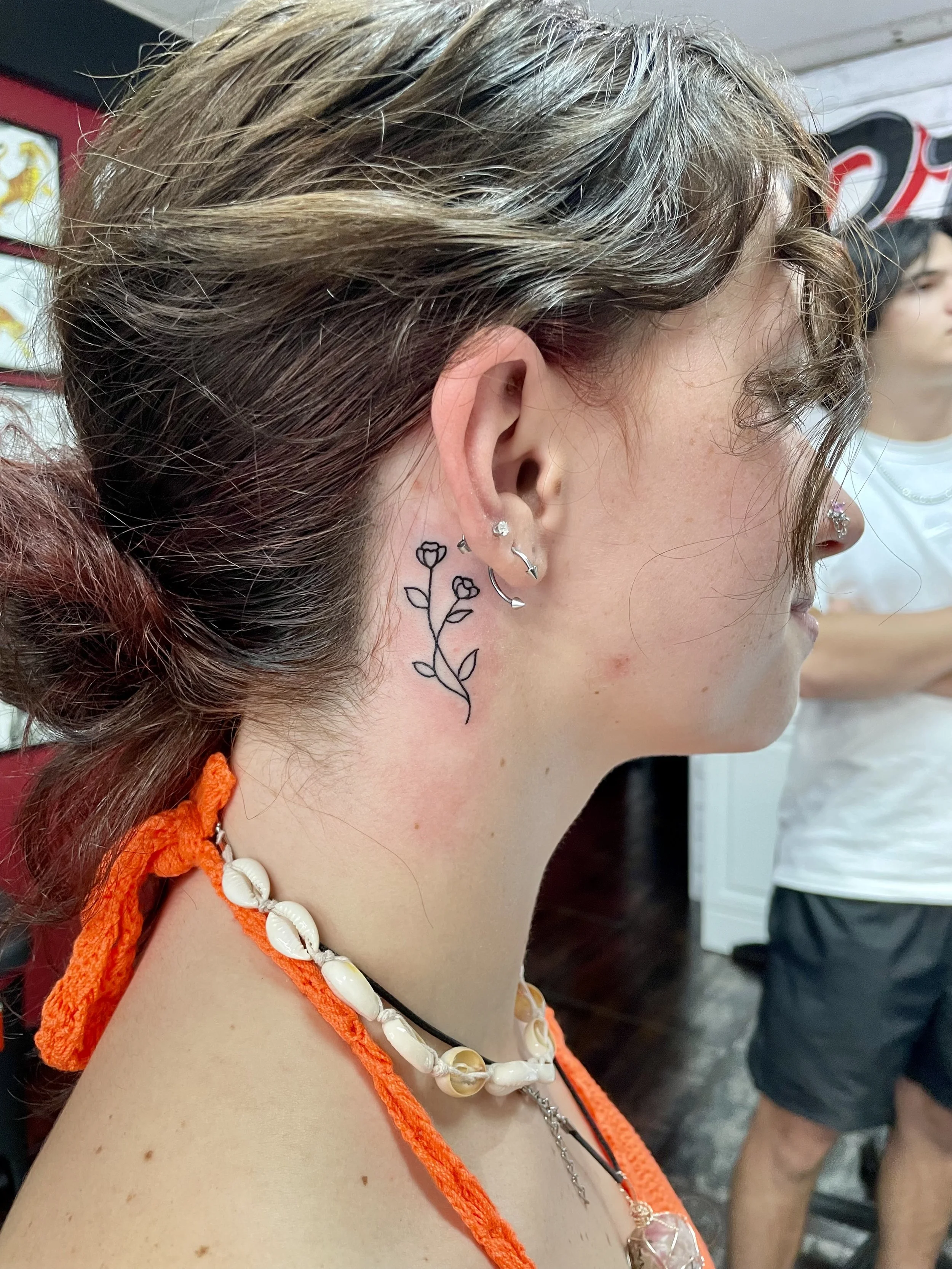 Fine line behind-the-ear neck tattoo featuring a slender floral stem with two small blooming roses and simple leaves, custom tattoo by Nick at Big Kahuna Tattoo in Boca Raton, FL.