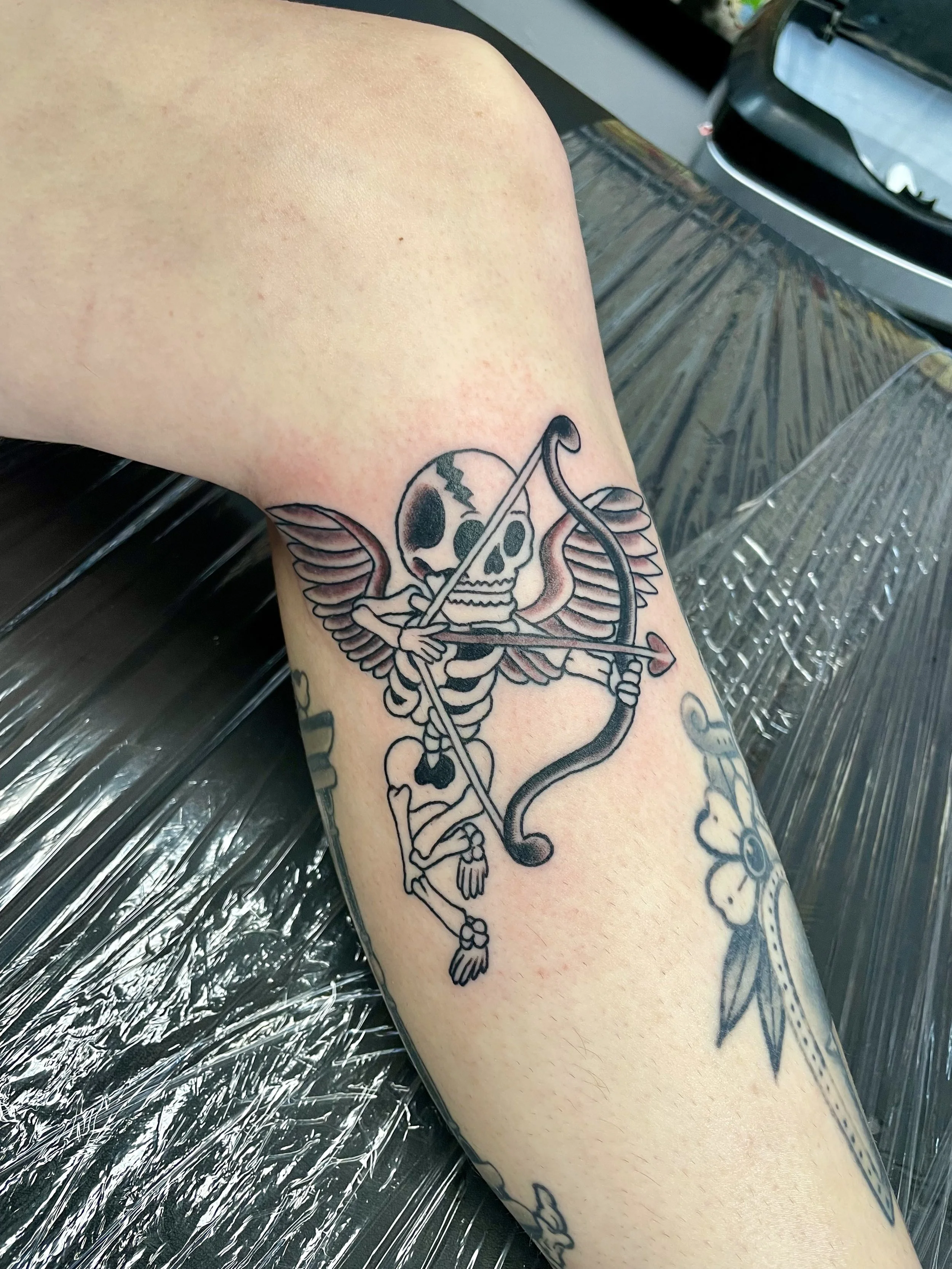 Black and grey forearm tattoo featuring a winged skeleton Cupid with a cracked skull, drawing back a bow aimed with a heart-tipped arrow, custom tattoo by Nick at Big Kahuna Tattoo in Boca Raton, FL.