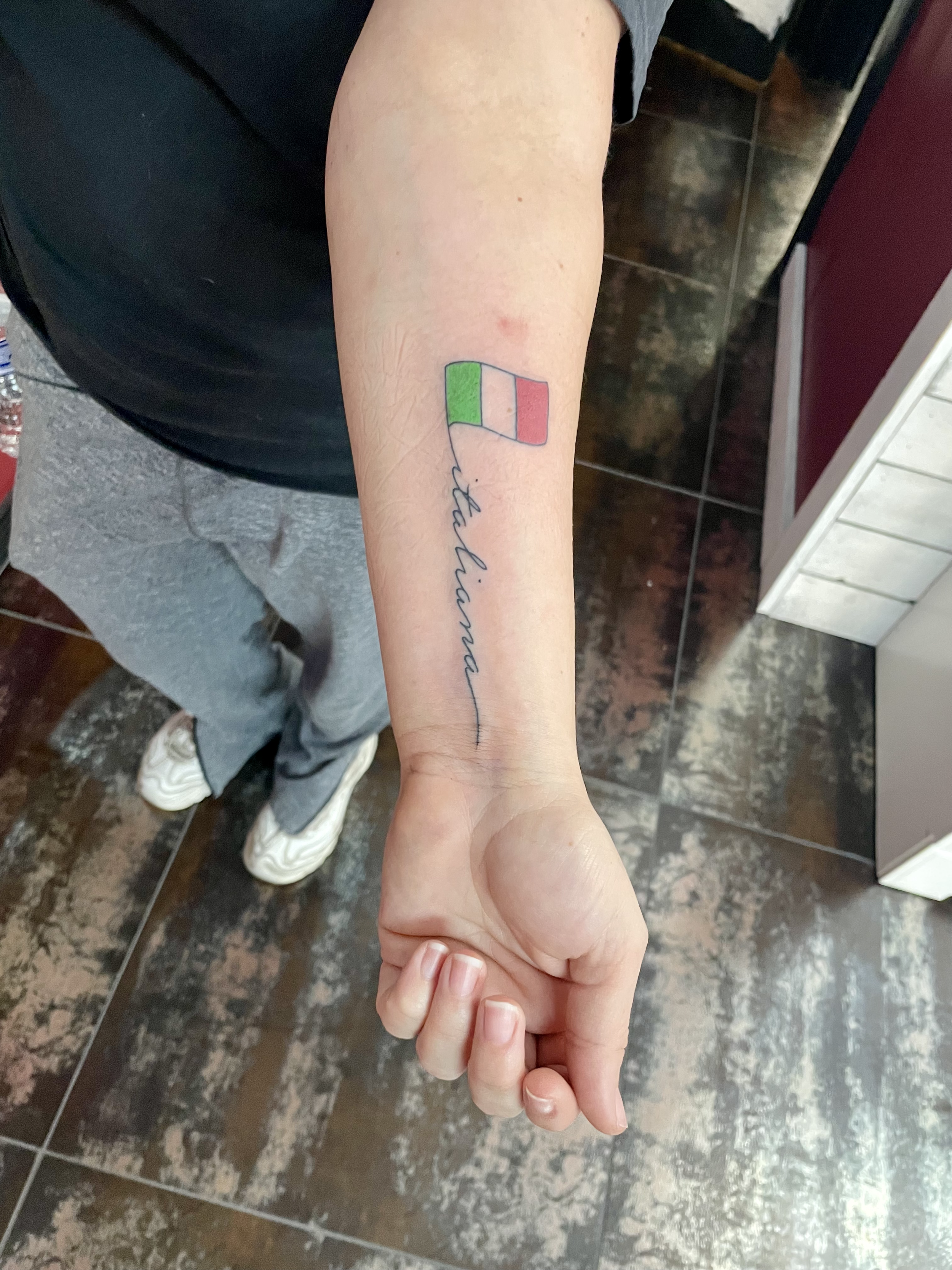 Fine line forearm tattoo featuring a small Italian flag with the word "italiana" in delicate cursive script, custom tattoo by Nick at Big Kahuna Tattoo in Boca Raton, FL.