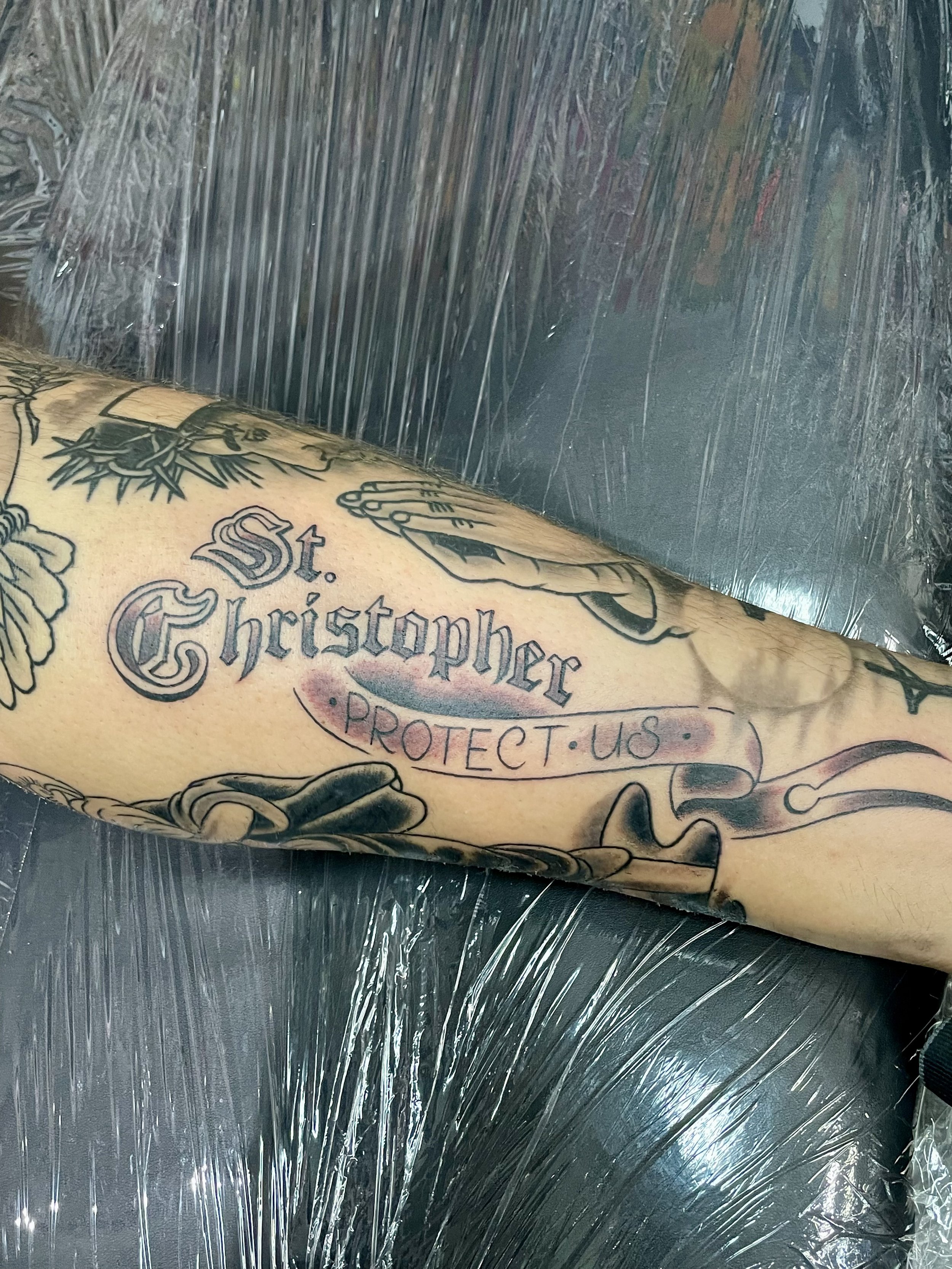 Black and grey forearm tattoo featuring bold Old English lettering reading "St. Christopher" above a flowing ribbon banner inscribed with "Protect Us," part of a larger religious themed sleeve, custom tattoo by Nick at Big Kahuna Tattoo in Boca Raton