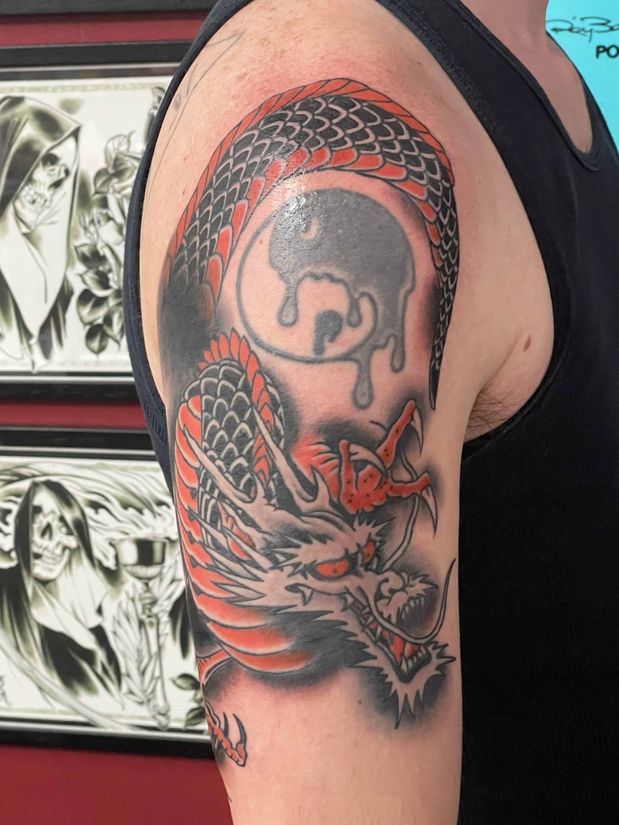Black scales with red accent plates and a snarling open jaw. This upper arm dragon coils around a preexisting dripping yin yang symbol, blending raw aggression with a deeper sense of balance. Japanese upper arm tattoo by Nick at Big Kahuna Tattoo in 