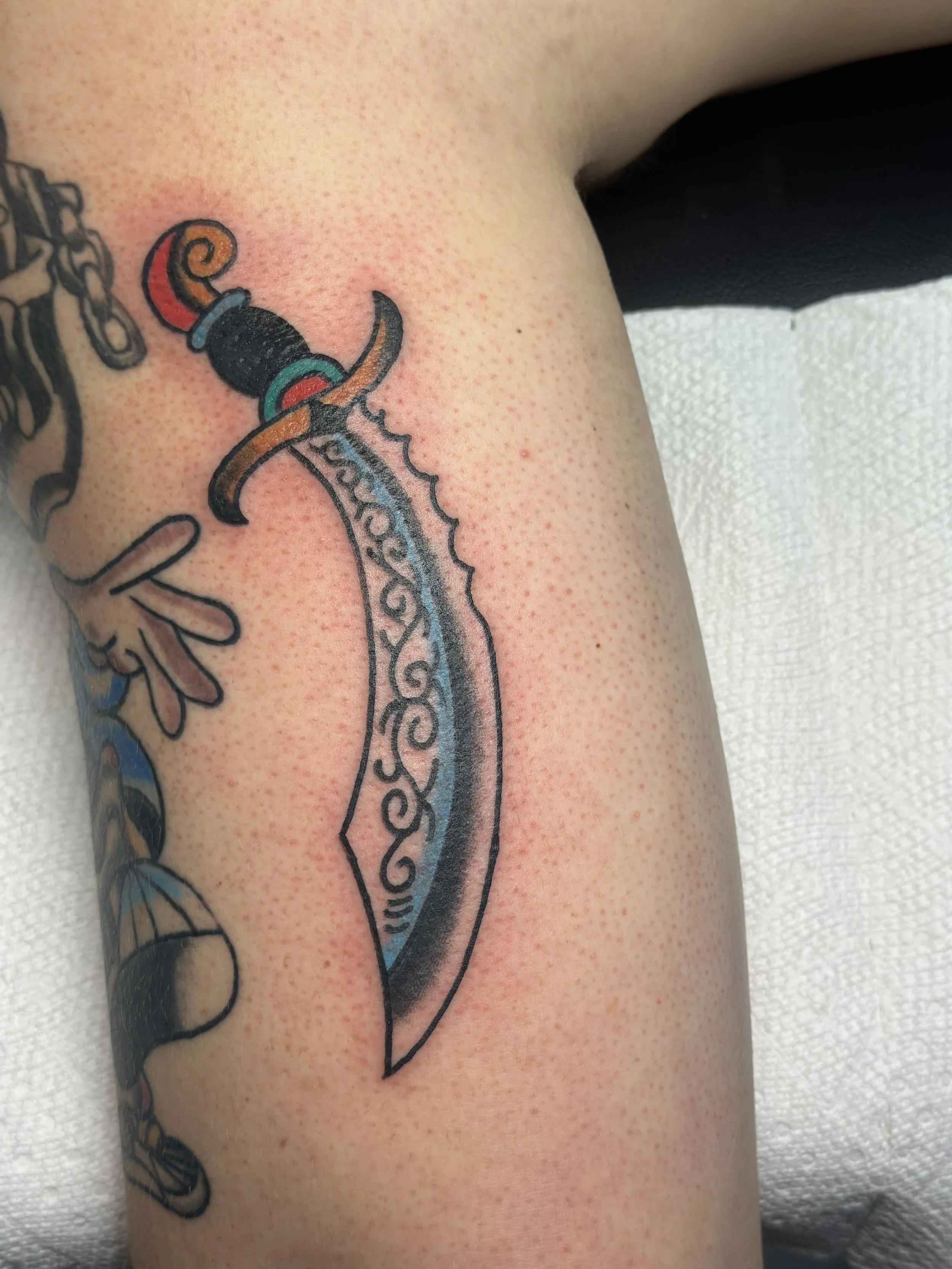 Traditional American color forearm tattoo featuring an ornate curved dagger with a blue blade etched with scroll detailing and a colorful red, teal, and gold hilt, custom tattoo by Dan at Big Kahuna Tattoo in Boca Raton, FL.