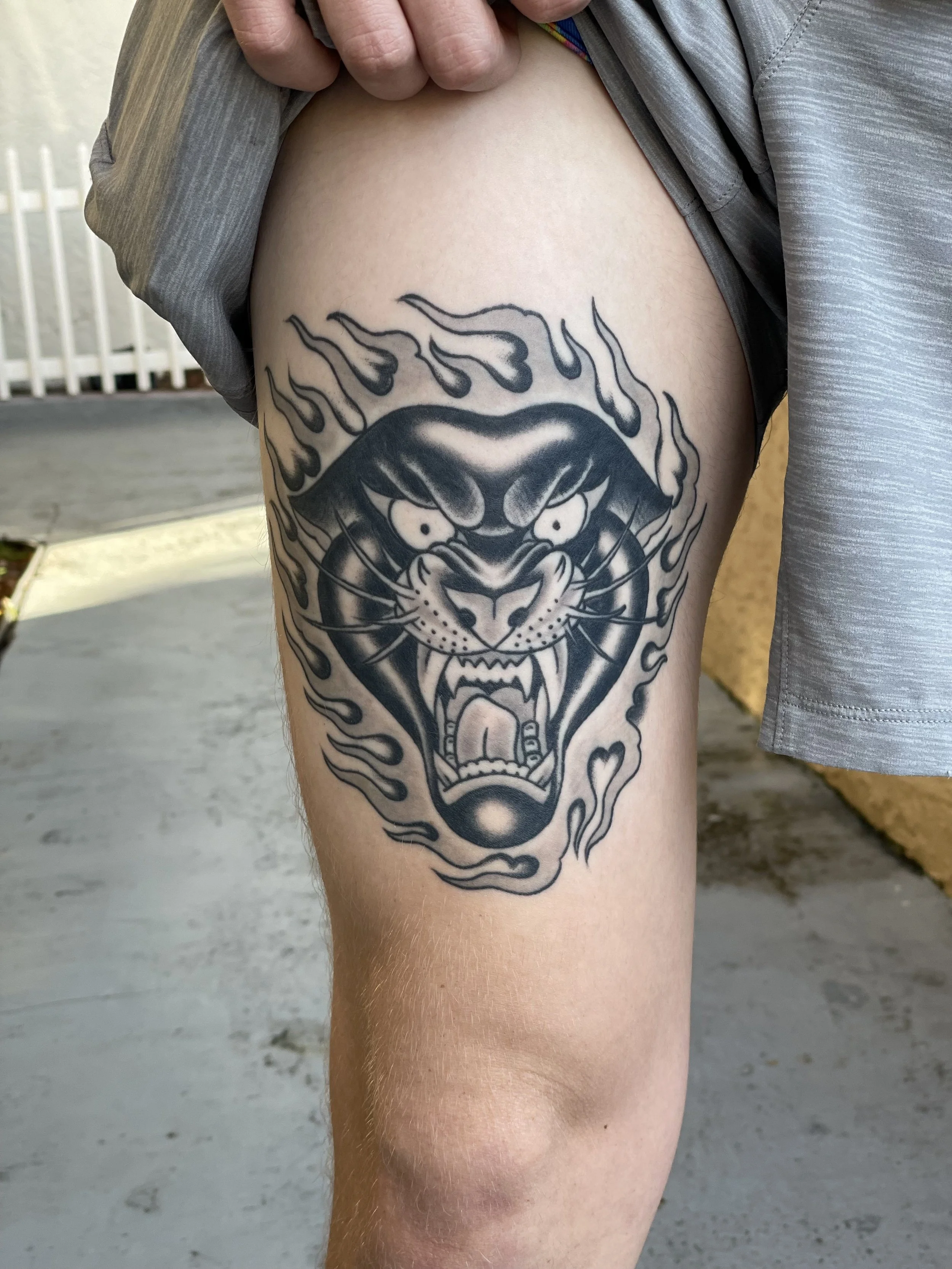 Pure attitude on the thigh. A snarling panther face front and center, surrounded by wild outlined flames that frame the whole tattoo in controlled chaos. Heavy black shading, no color needed. Black and grey thigh tattoo by Nick at Big Kahuna Tattoo i