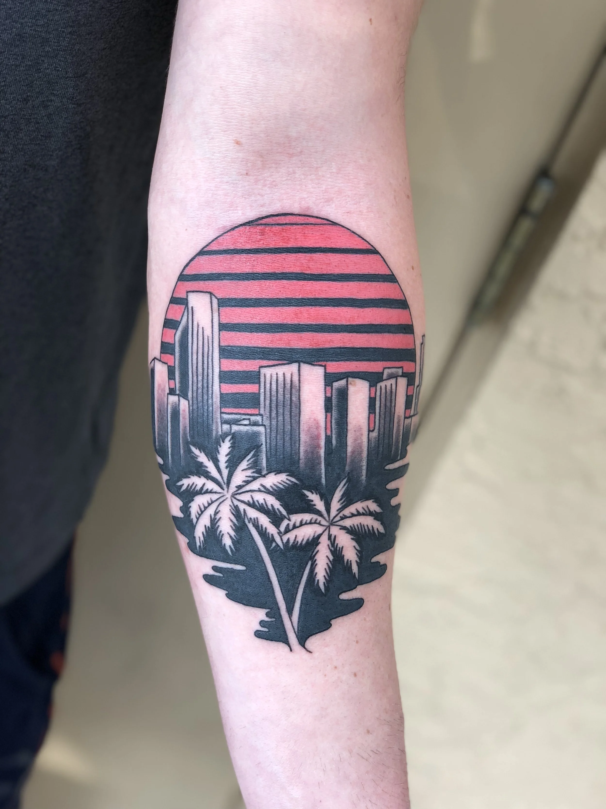 Bold graphic forearm tattoo featuring a retro Miami style cityscape with skyscrapers, palm trees, and a striped red sunset in black and red, custom tattoo by Nick at Big Kahuna Tattoo in Boca Raton, FL.